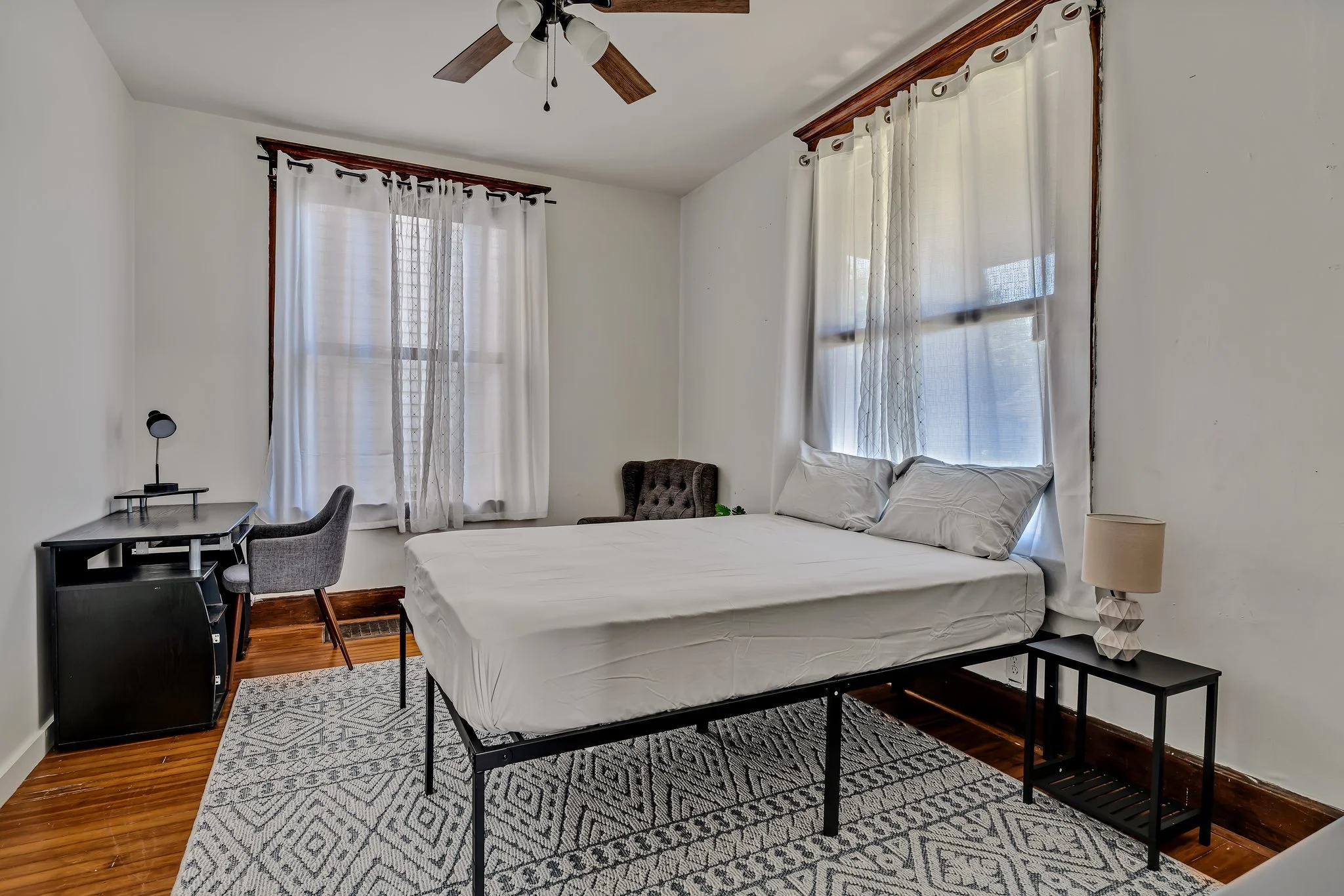 A bedroom with two large windows with white curtains, a bed with white sheets and pillows, a nightstand with a lamp, a small desk with a chair and a black mini fridge, and a brown armchair in the corner. The room has hardwood floors, a patterned rug, and a ceiling fan.