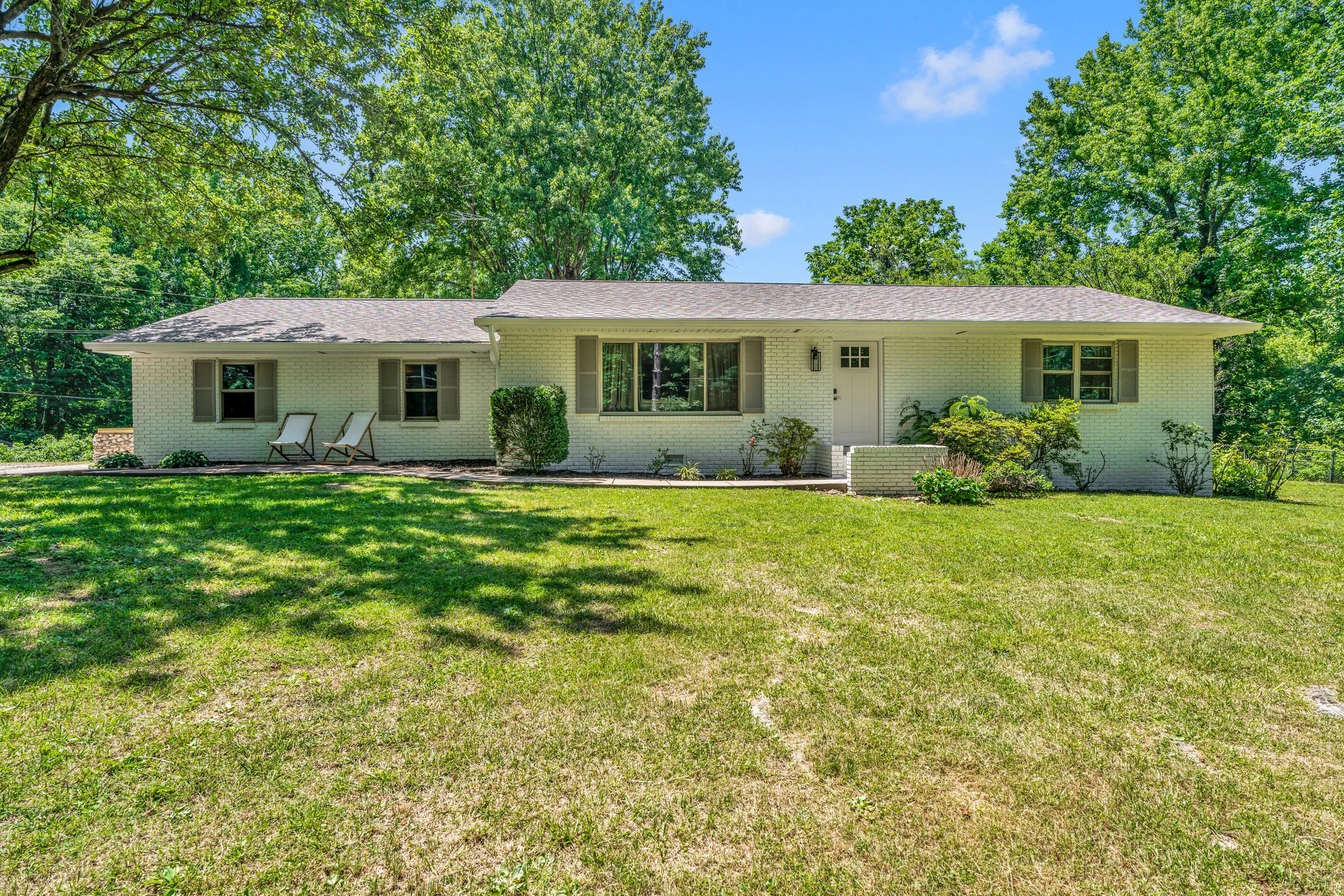 A single-story house with white brick exterior, gray shingle roof, surrounded by green trees and a well-maintained lawn, with two chairs on the front porch.