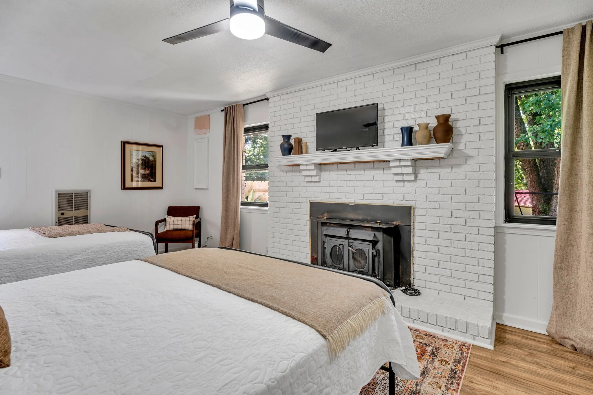 A room with two beds, a white brick fireplace, a mounted TV above the fireplace, and a shelf with decorative vases. There are two windows with curtains, a ceiling fan with lights, and a framed picture on the wall.