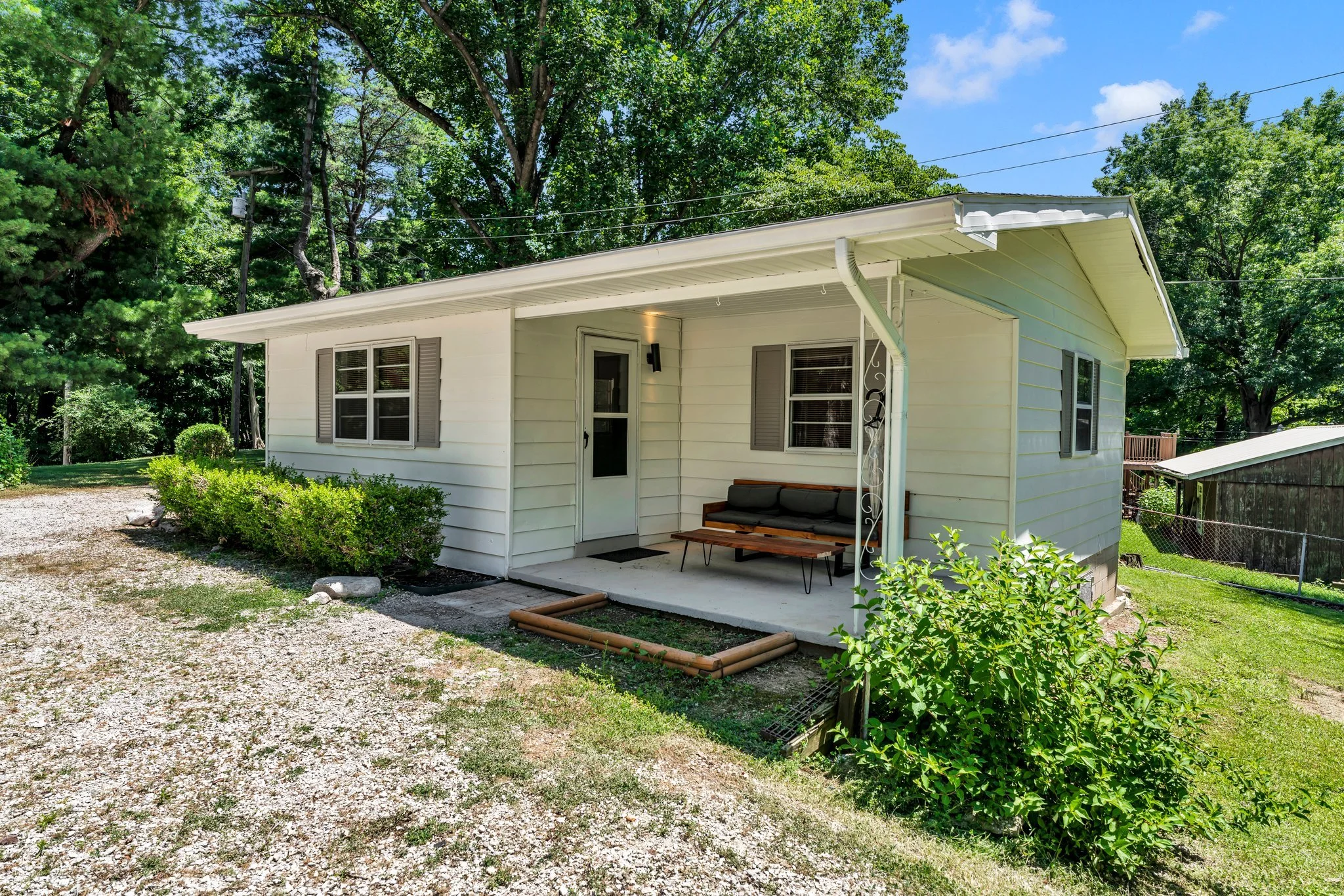 Small white house with a covered front porch, two windows with shutters, and lush green trees in the background.