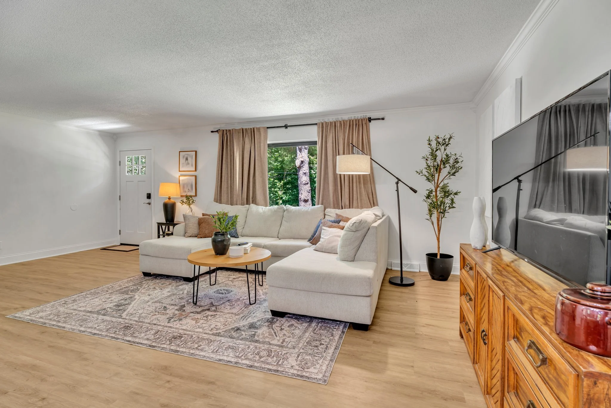 Living room with white sectional sofa, beige curtains, wooden floor, TV on wooden console, potted plants, and decorative lamps.