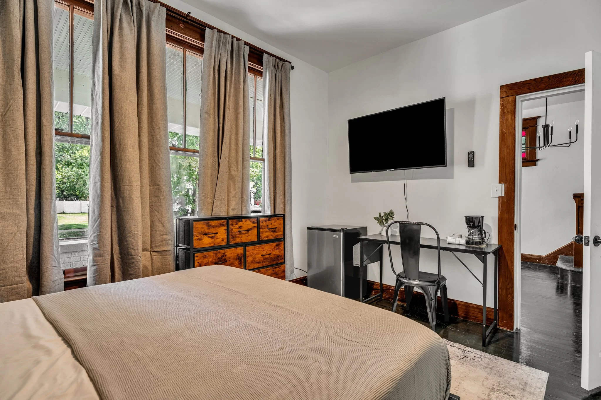 A bedroom with beige bedding, large windows with beige curtains, a small dresser, a wall-mounted TV, a desk with a chair, a coffee maker, and a mini fridge, with a view of a porch outside.