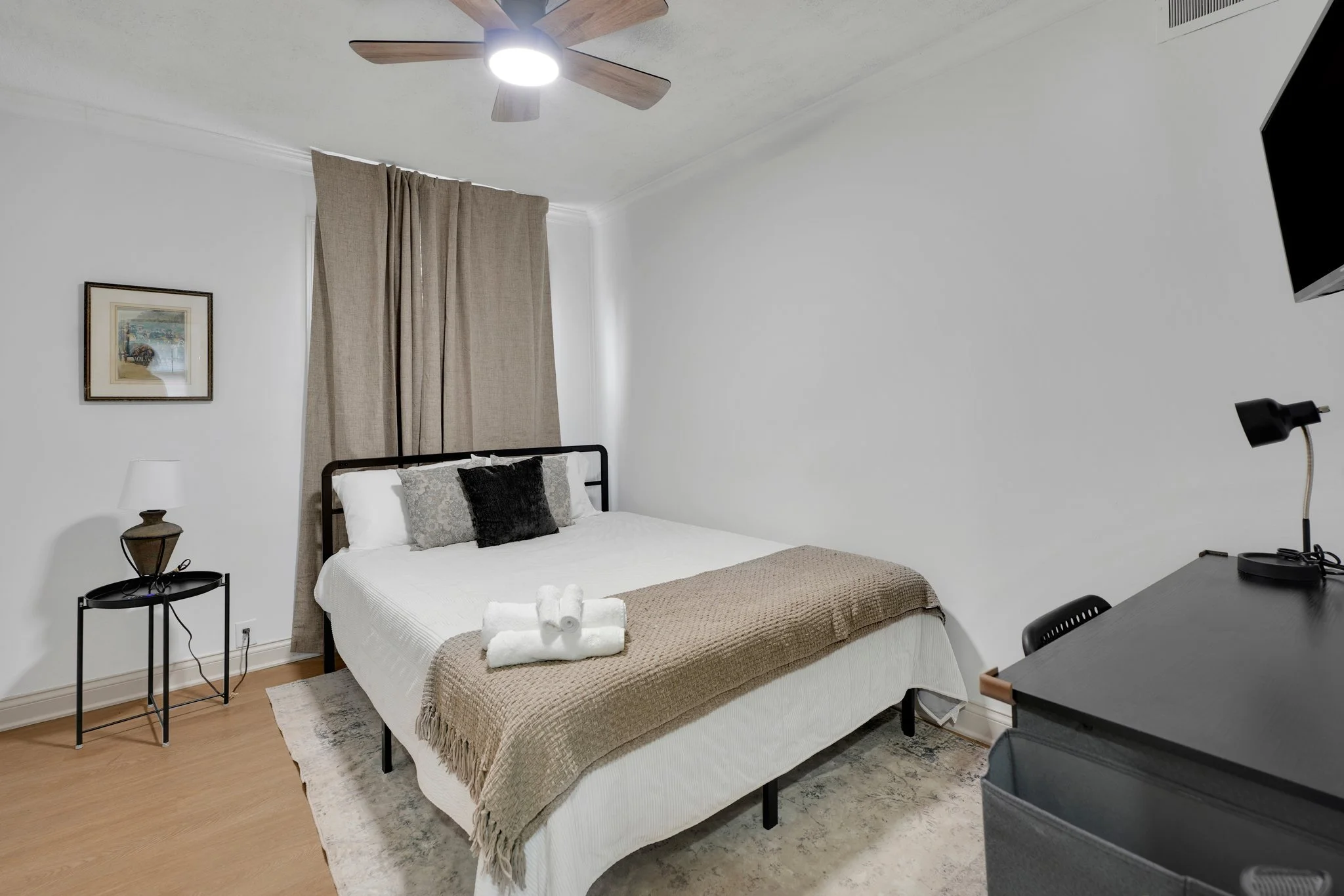 A neatly made bedroom featuring a bed with white linens, decorative pillows, and a beige throw blanket, topped with rolled towels. To the left, a black side table with a lamp. Behind the bed, beige curtains cover a window, with a framed picture hanging on the white wall. A ceiling fan with wooden blades is mounted on the ceiling. To the right, there's a black desk with a black lamp and a wall-mounted flat-screen TV.