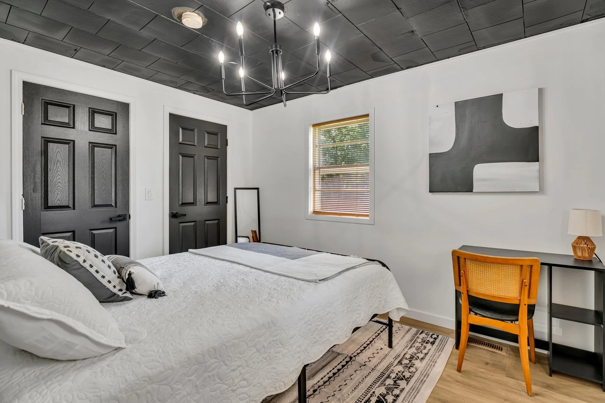 A bedroom with a white bed, black doors, a window with blinds, a black ceiling with a chandelier, a painting on the wall, a mirror, a chair, and a desk with a lamp.