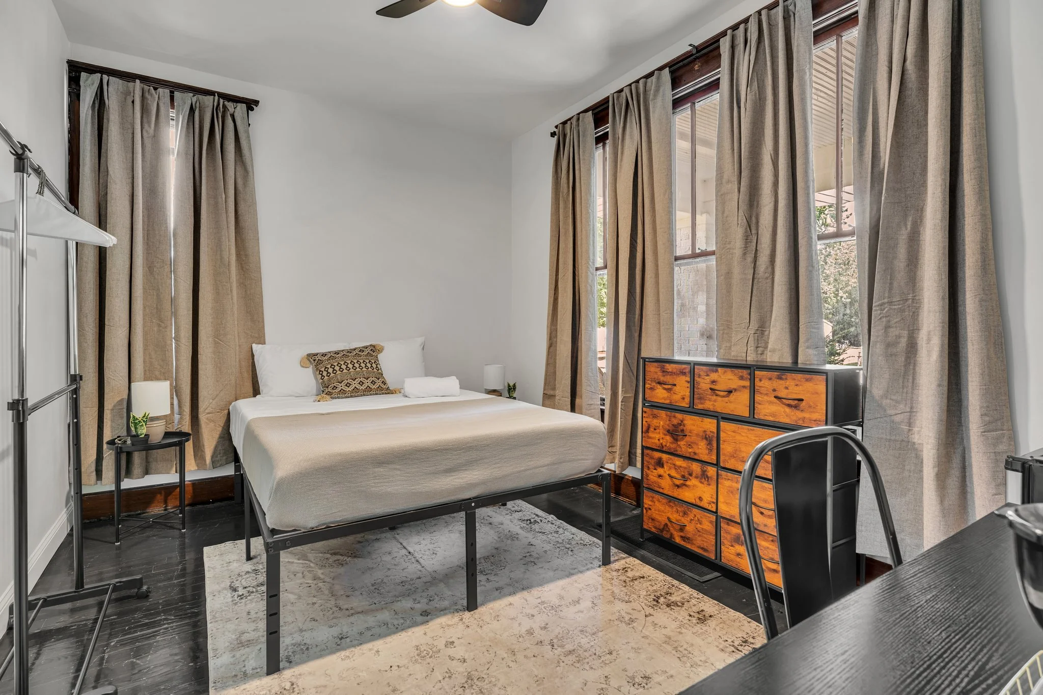 Bedroom with a bed, beige bedding, decorative pillow, nightstands with lamps, large windows with beige curtains, wooden dresser, black dining chair, black table, and a rug.