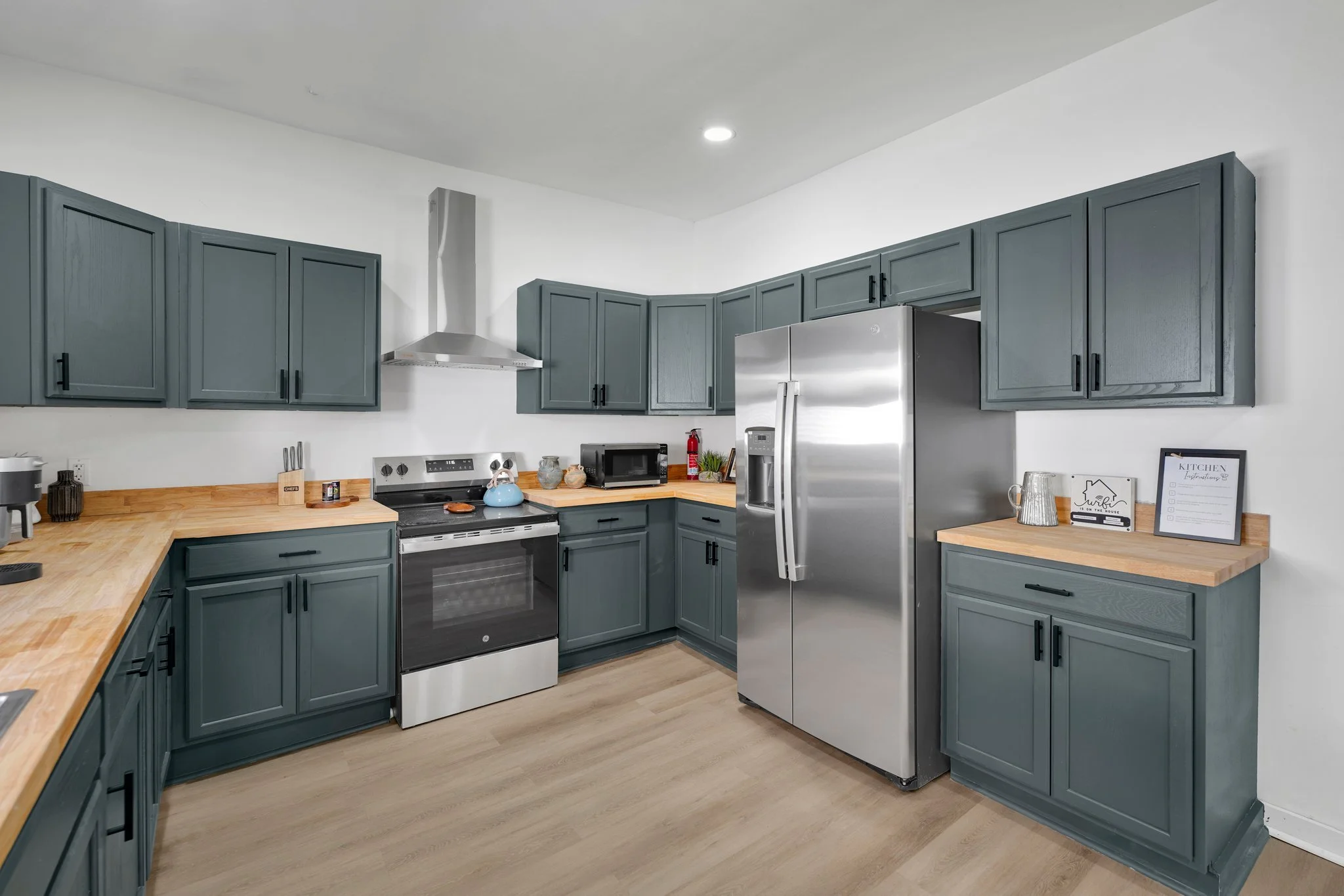 Modern kitchen with blue cabinets, wooden countertops, stainless steel appliances including a refrigerator, oven, and microwave, and light-colored wood flooring.