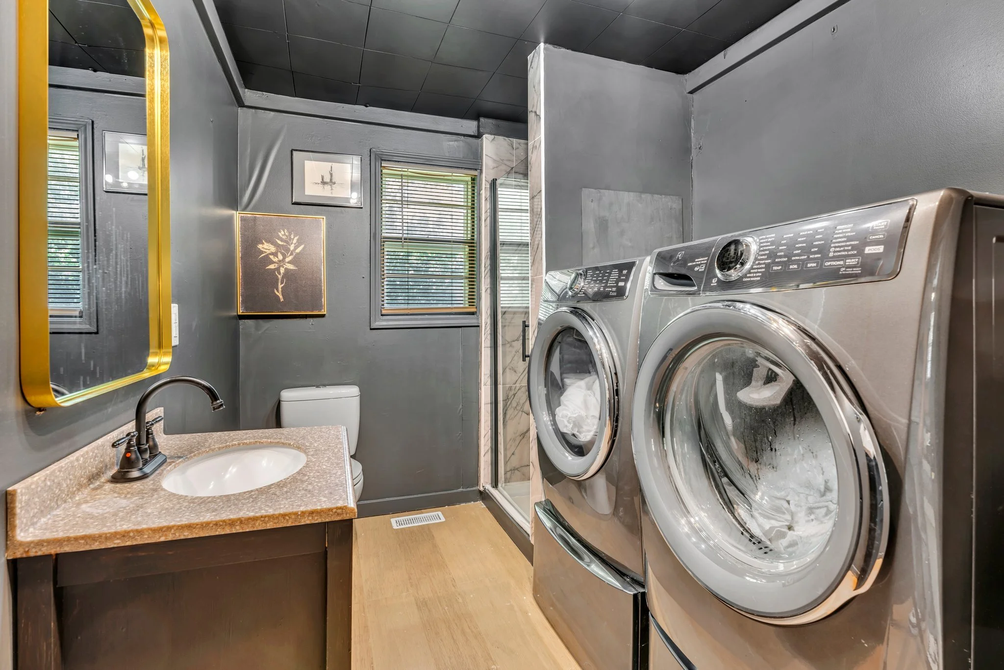 Laundry room with front-loading washer and dryer, a small sink with a beige countertop, a toilet, framed artwork, windows with blinds, a mirror with a gold frame, and a shower area with a glass door.