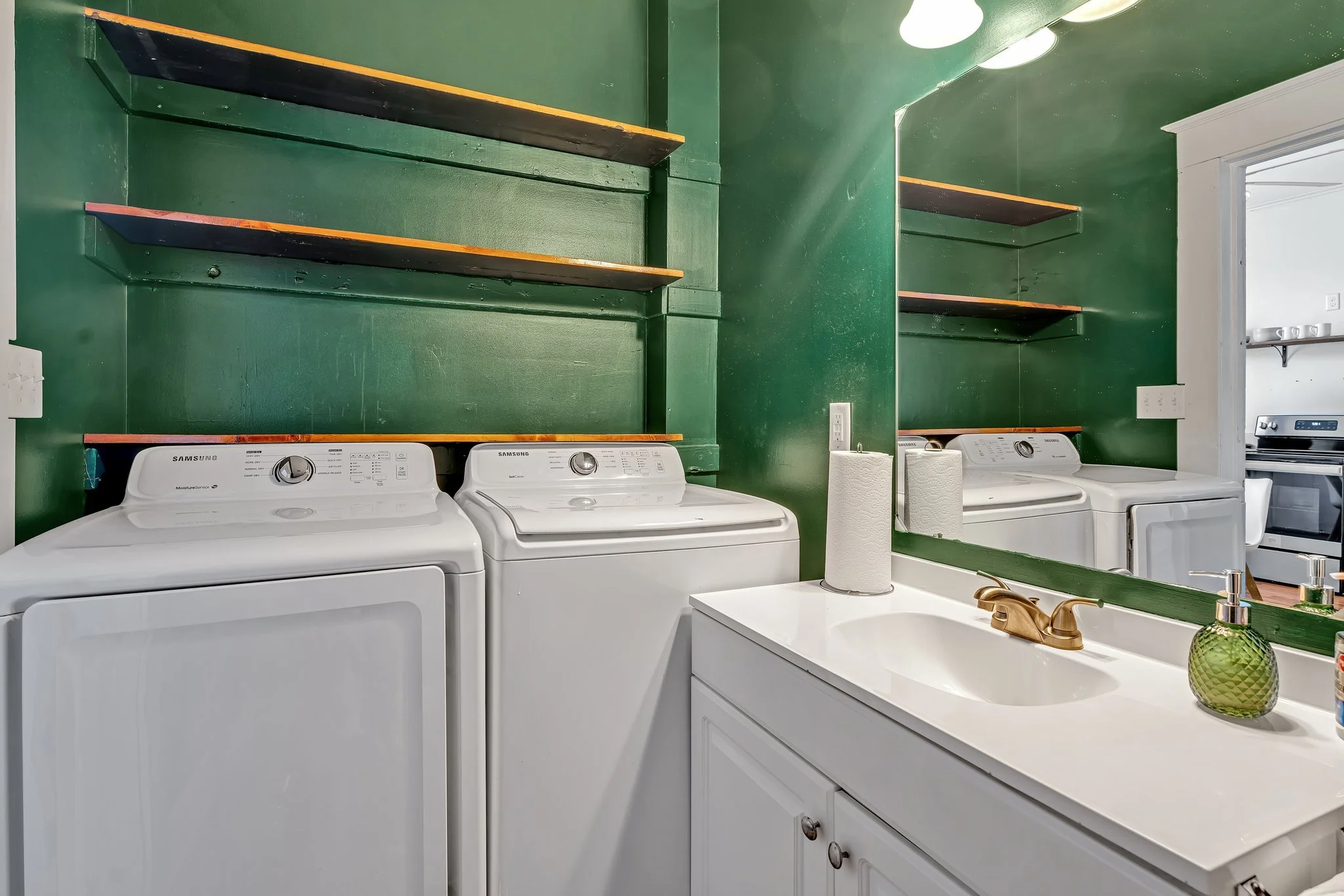 Laundry room with green walls, shelves, white washer and dryer, sink, mirror, paper towels, soap dispenser, and a view into another room with a stove.