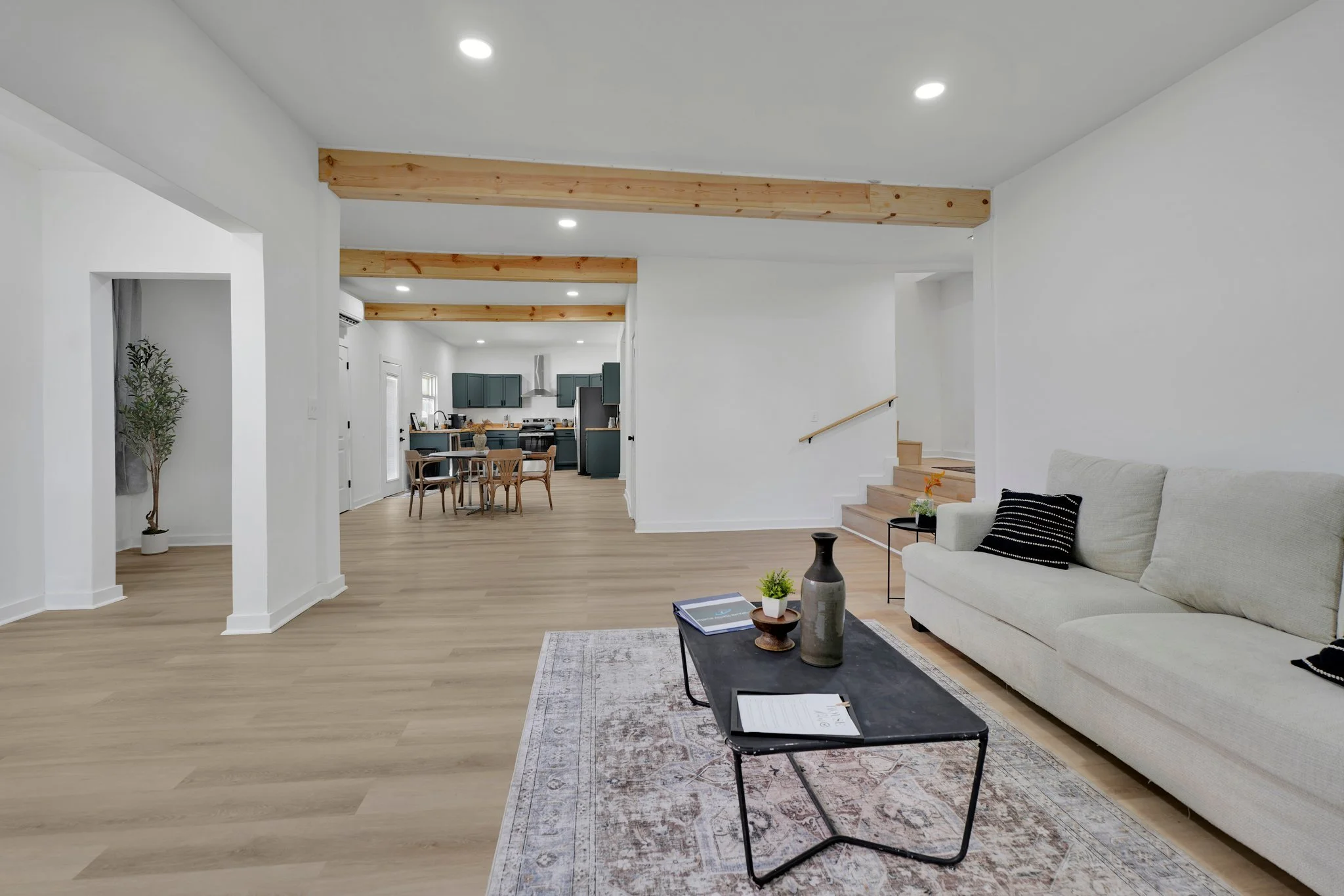 Living room with beige sofa, black coffee table with decorative vases and books, rug, and wooden floors, open to the dining area and kitchen with teal cabinets, wooden beams on ceiling, and staircase with wooden steps.