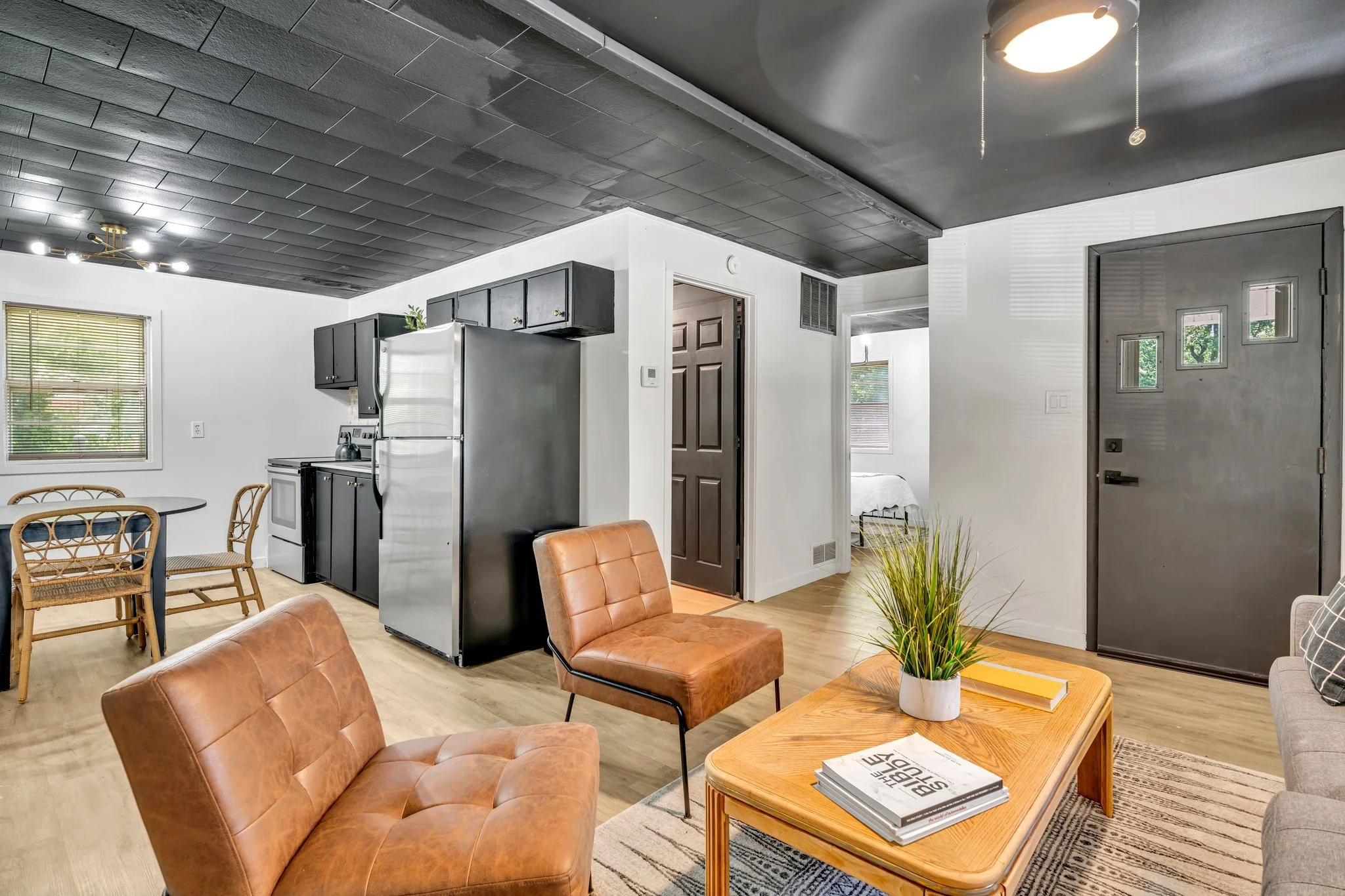 Living room with leather chairs, wooden coffee table, potted plant, and a backdrop of kitchen and doorway leading to a bedroom. Walls are white, ceiling is black, with modern lighting, and hardwood flooring.