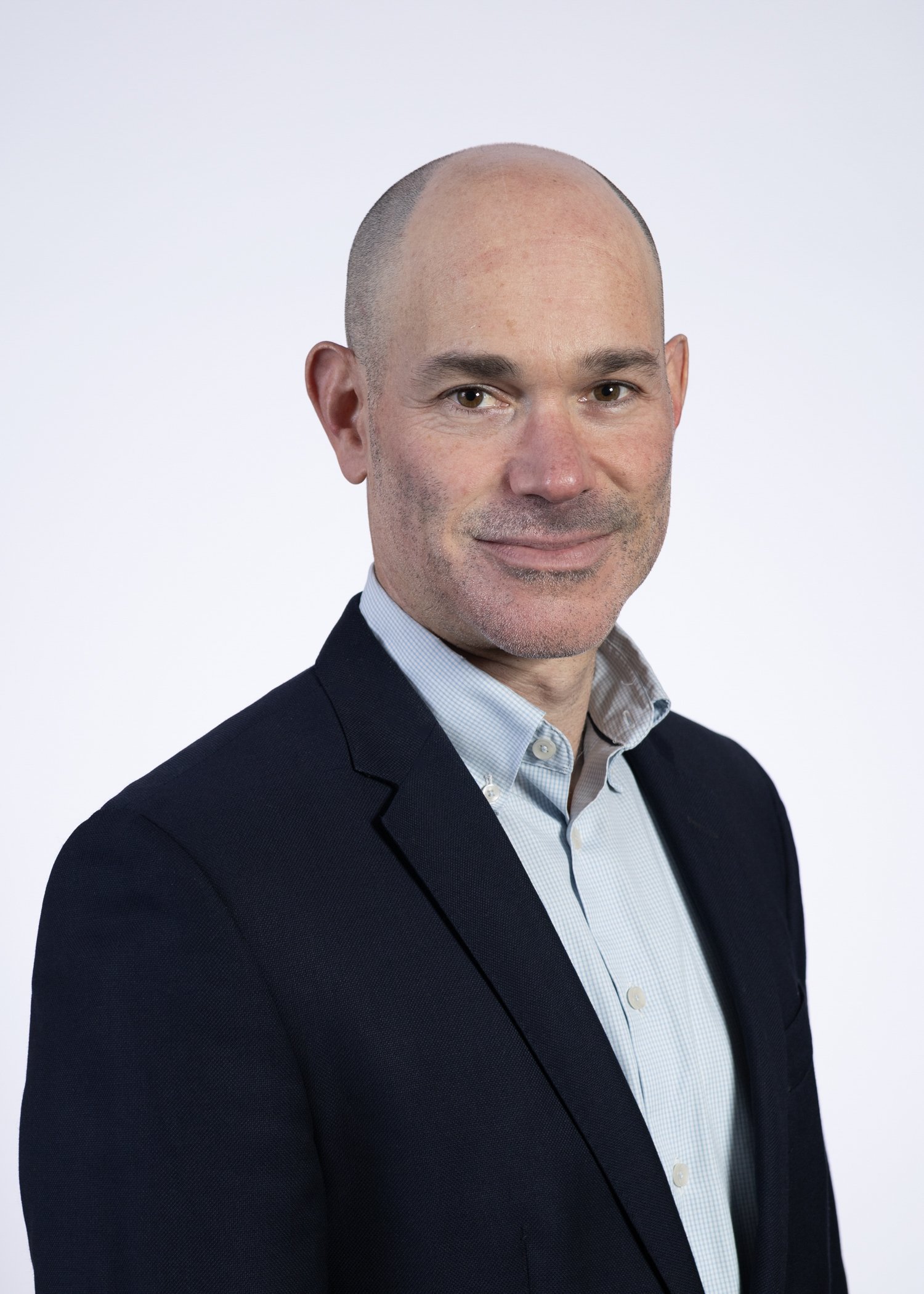 A professional headshot of a bald man in a dark suit jacket and light-colored dress shirt, standing against a plain white background.