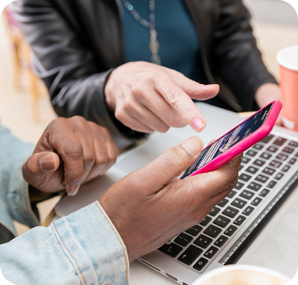 Two people looking at a smartphone together over a laptop, with coffee cups nearby.
