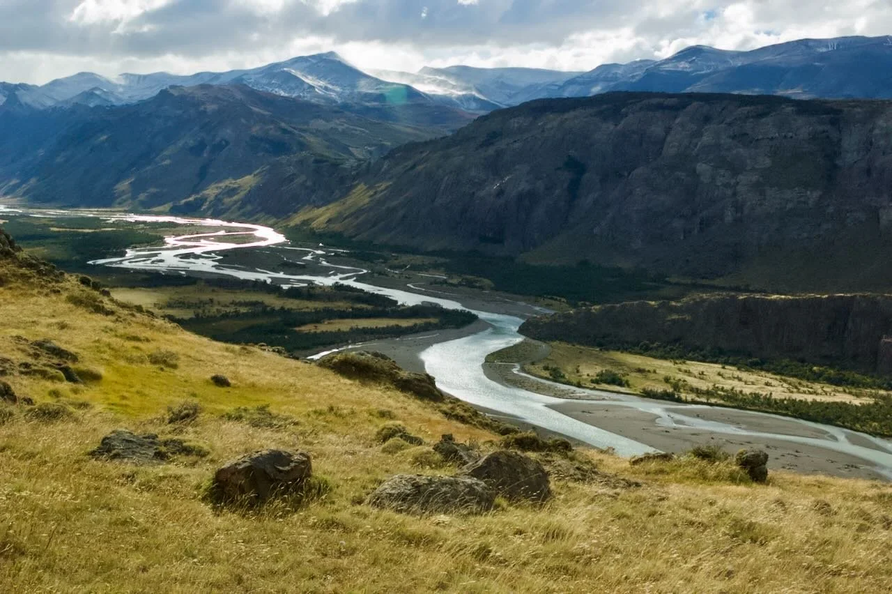Scenic view of a winding river flowing through a valley with grassy hills and towering mountains in the background under a partly cloudy sky.