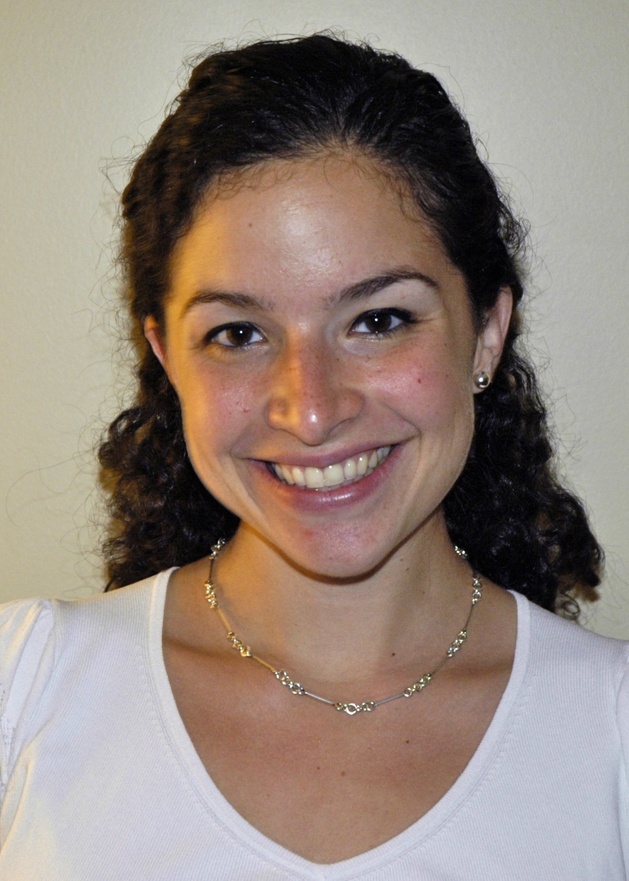 A woman with curly dark hair smiling, wearing a white top, a gold necklace, and earrings, against a plain light-colored background.