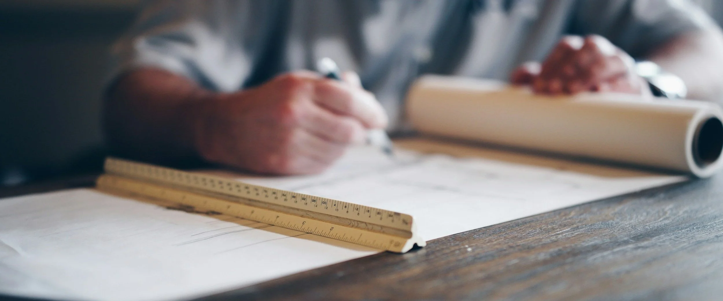 Person's hand writing on architectural plans with a ruler nearby and a rolled-up blueprint on the table.