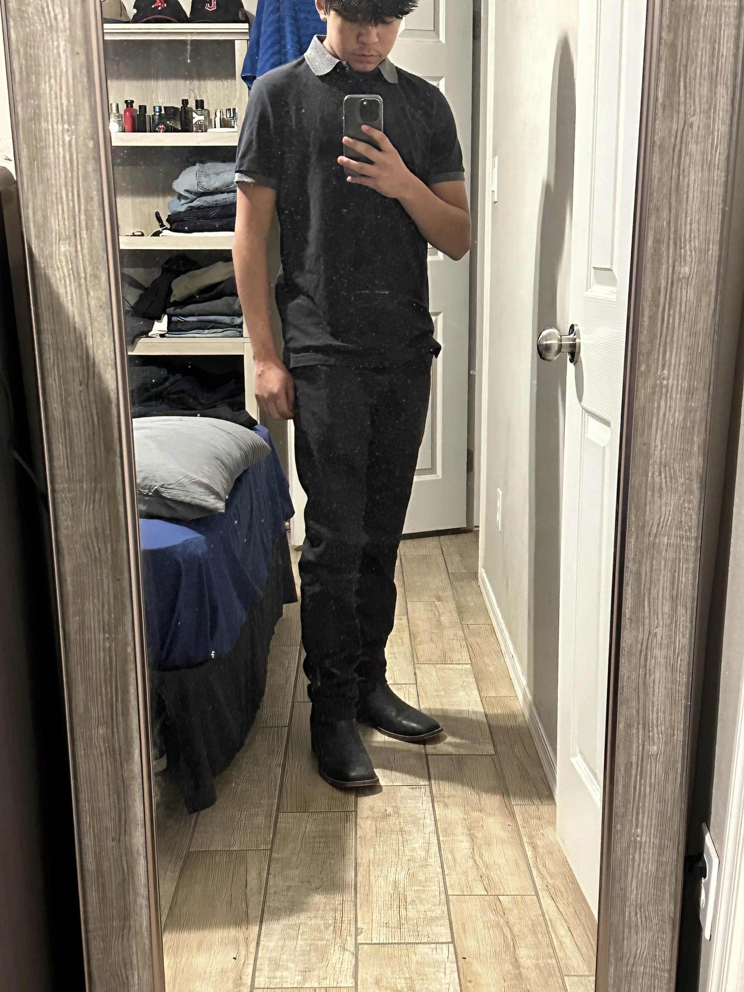 A person taking a mirror selfie in a bedroom, wearing a black polo shirt and black pants with cowboy boots.