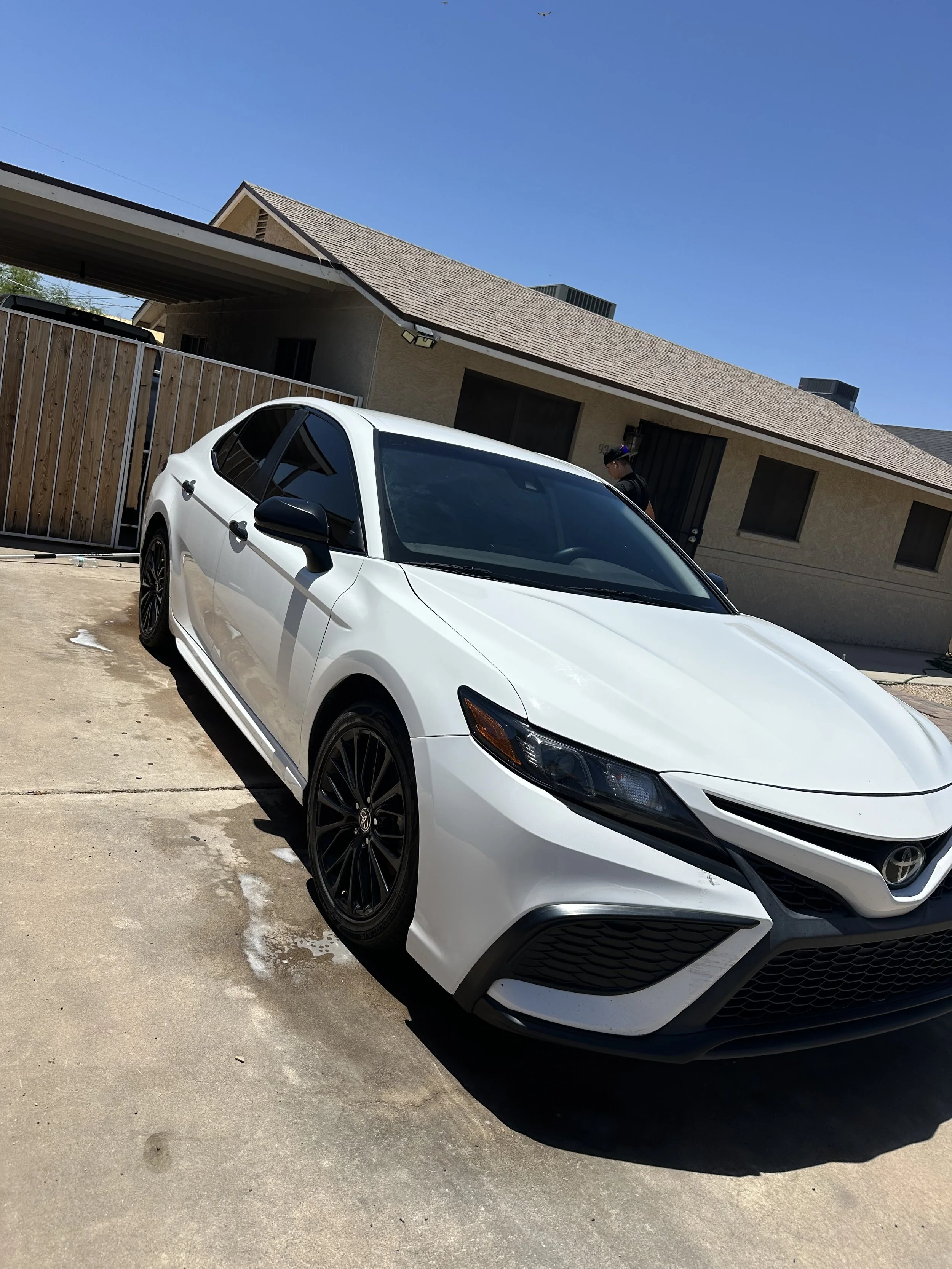 White Toyota sedan parked on a driveway in front of a house with a wooden fence and a clear blue sky.