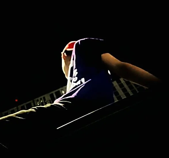 A person sitting outdoors at night, wearing a hooded jacket, facing away from the camera, with a building in the background.