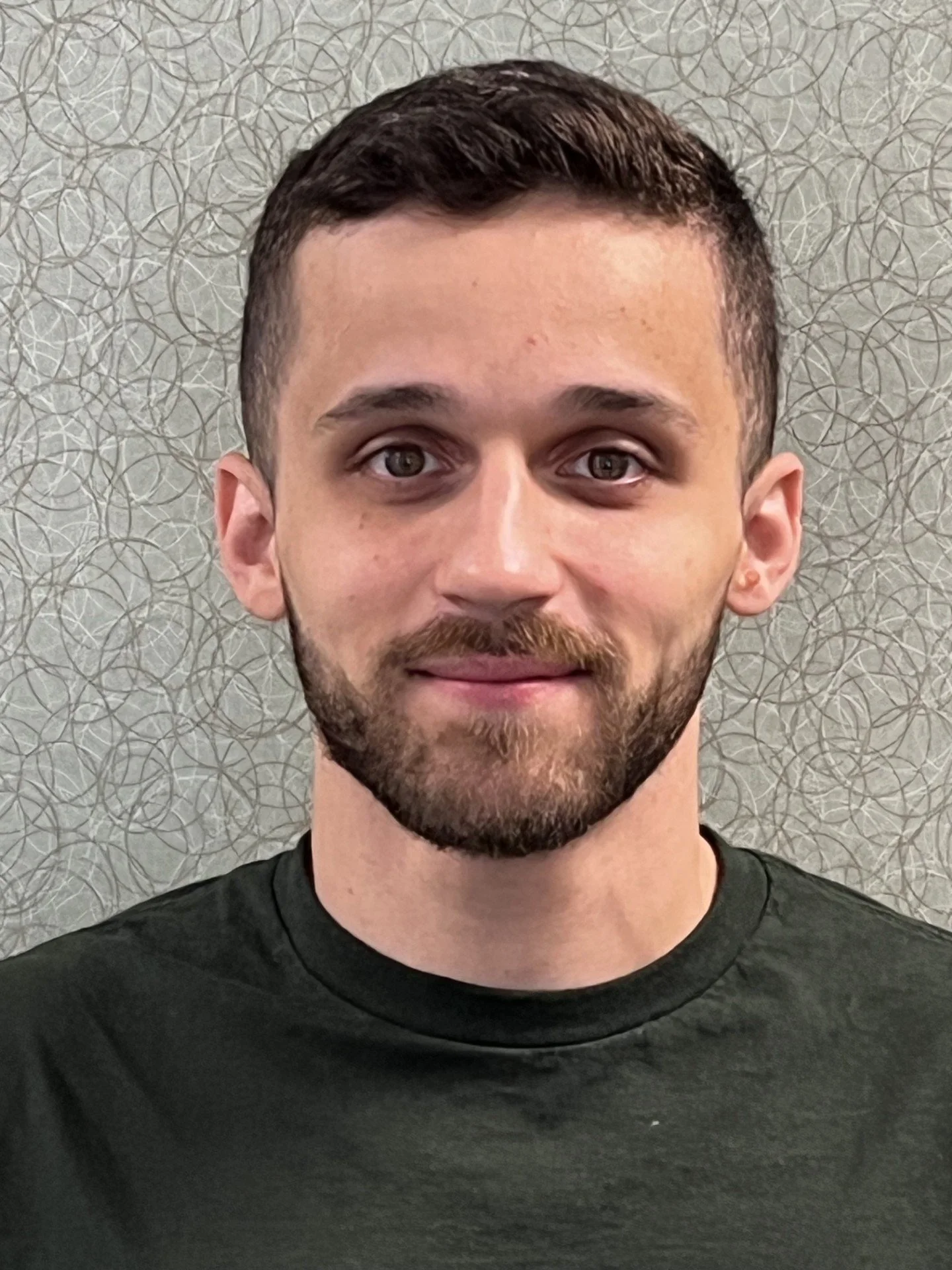 A smiling young man with dark hair and a beard, wearing a green shirt, standing in front of a textured green patterned background.