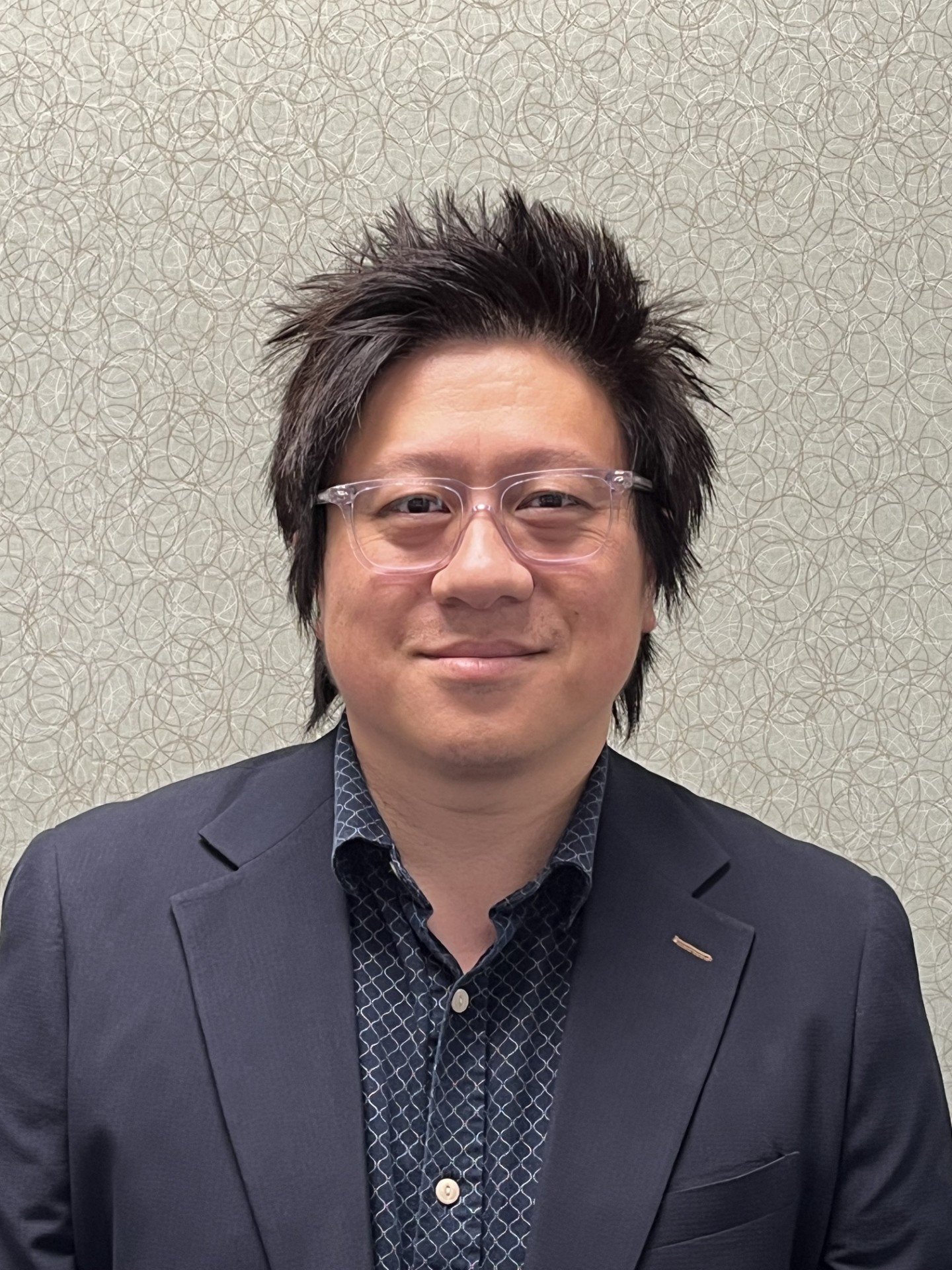 A man wearing glasses, a patterned dress shirt, and a dark blazer, smiling in front of a textured green wall.