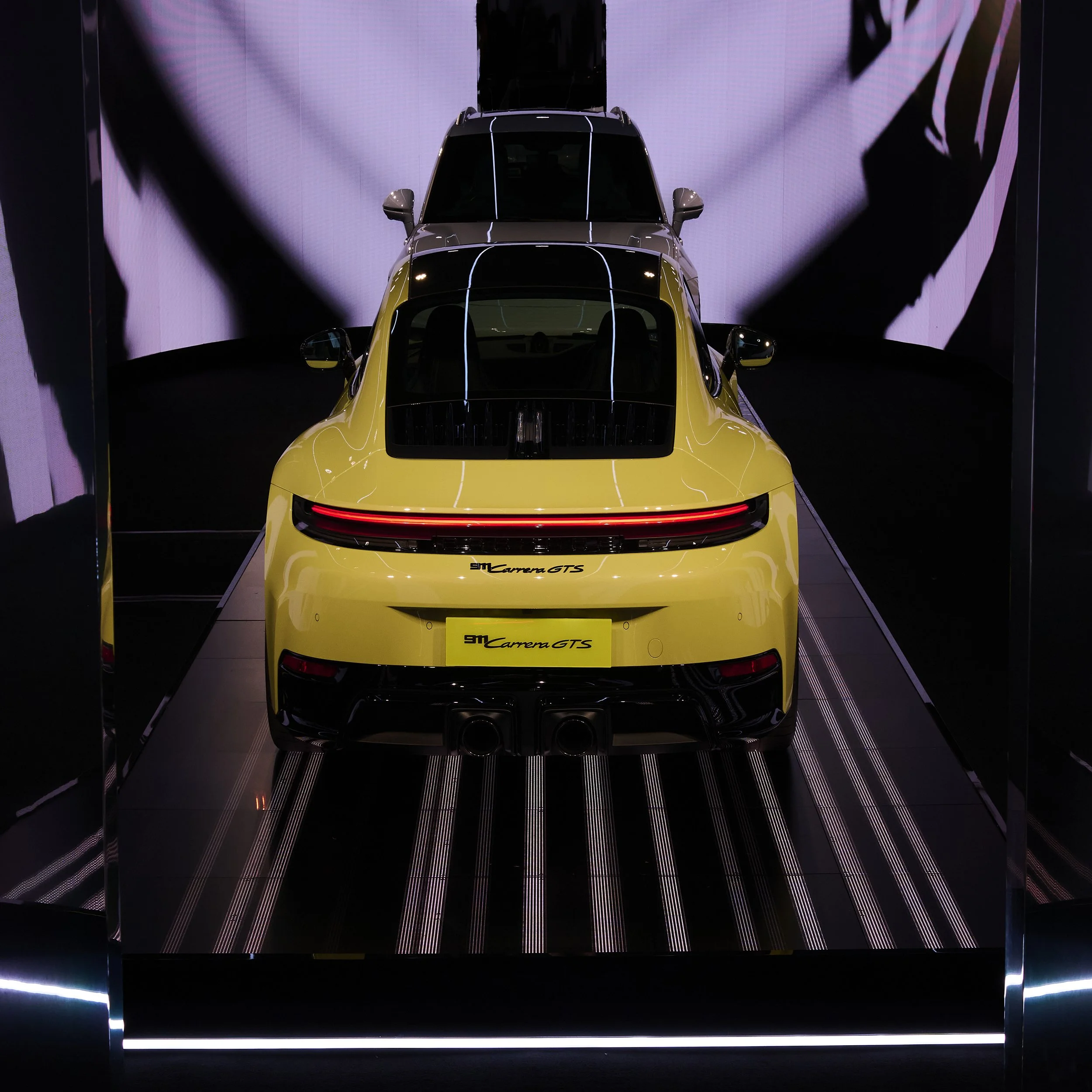 Yellow and black Porsche 911 Carrera GTS on display in a showroom with dark walls and striped flooring.