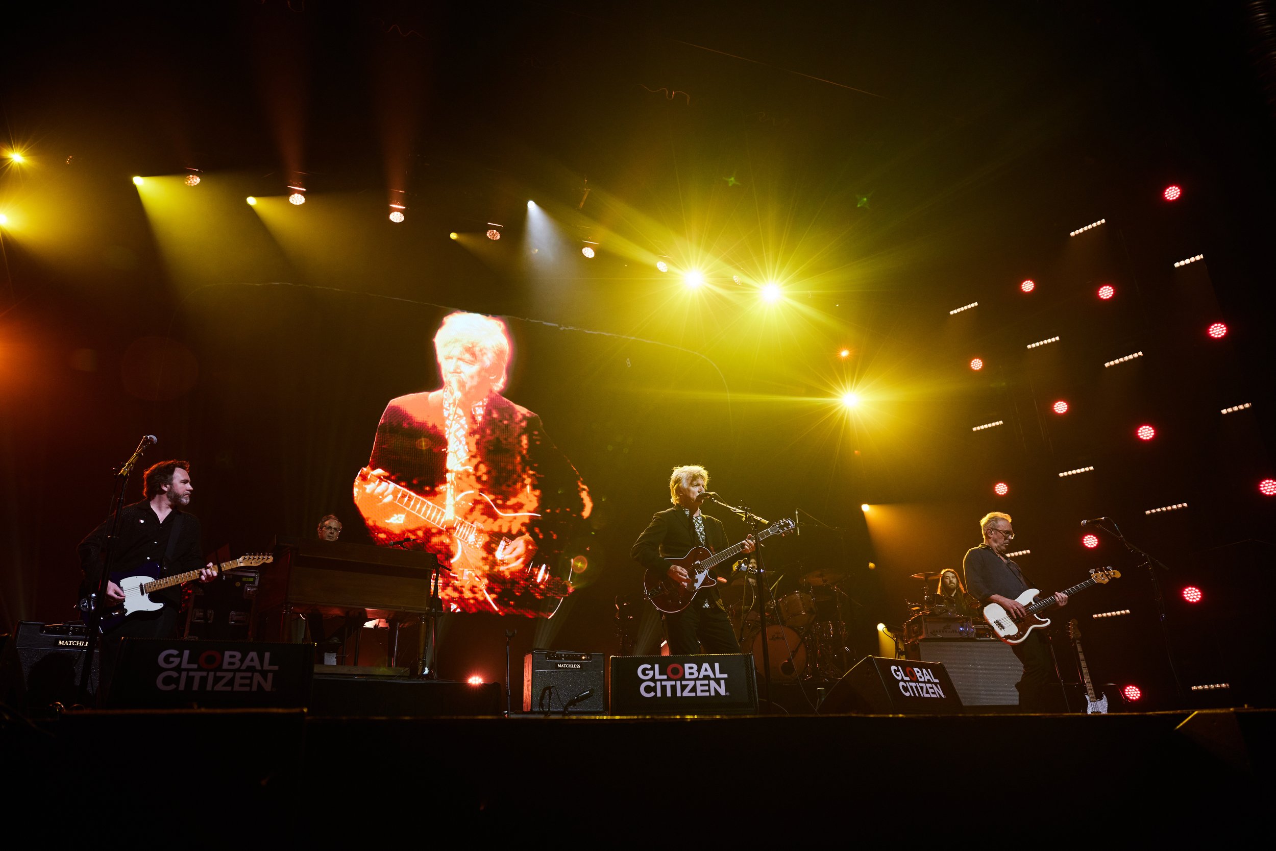 A band performing on stage at a Global Citizen event, with bright stage lighting and large screen projections of a guitarist. Musicians are playing guitars and keyboards with "Global Citizen" signs on stage.