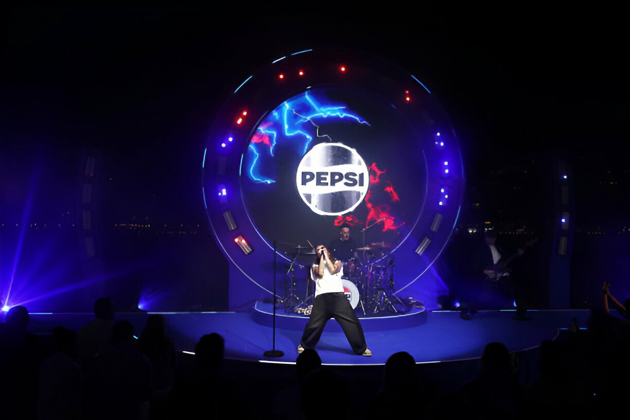 Concert stage with a singer performing in front of a large Pepsi logo, surrounded by colorful lights and audience.