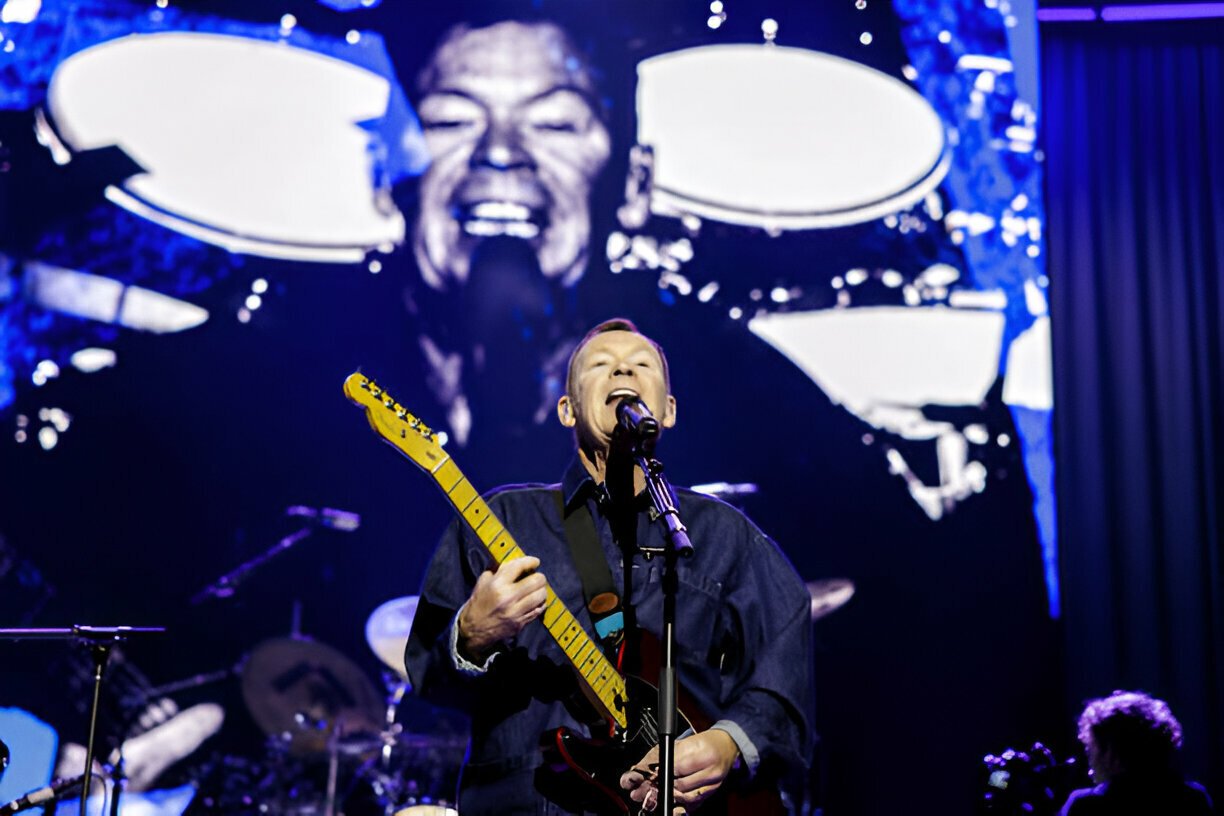 Musician performing on stage with electric guitar, large video screen backdrop.