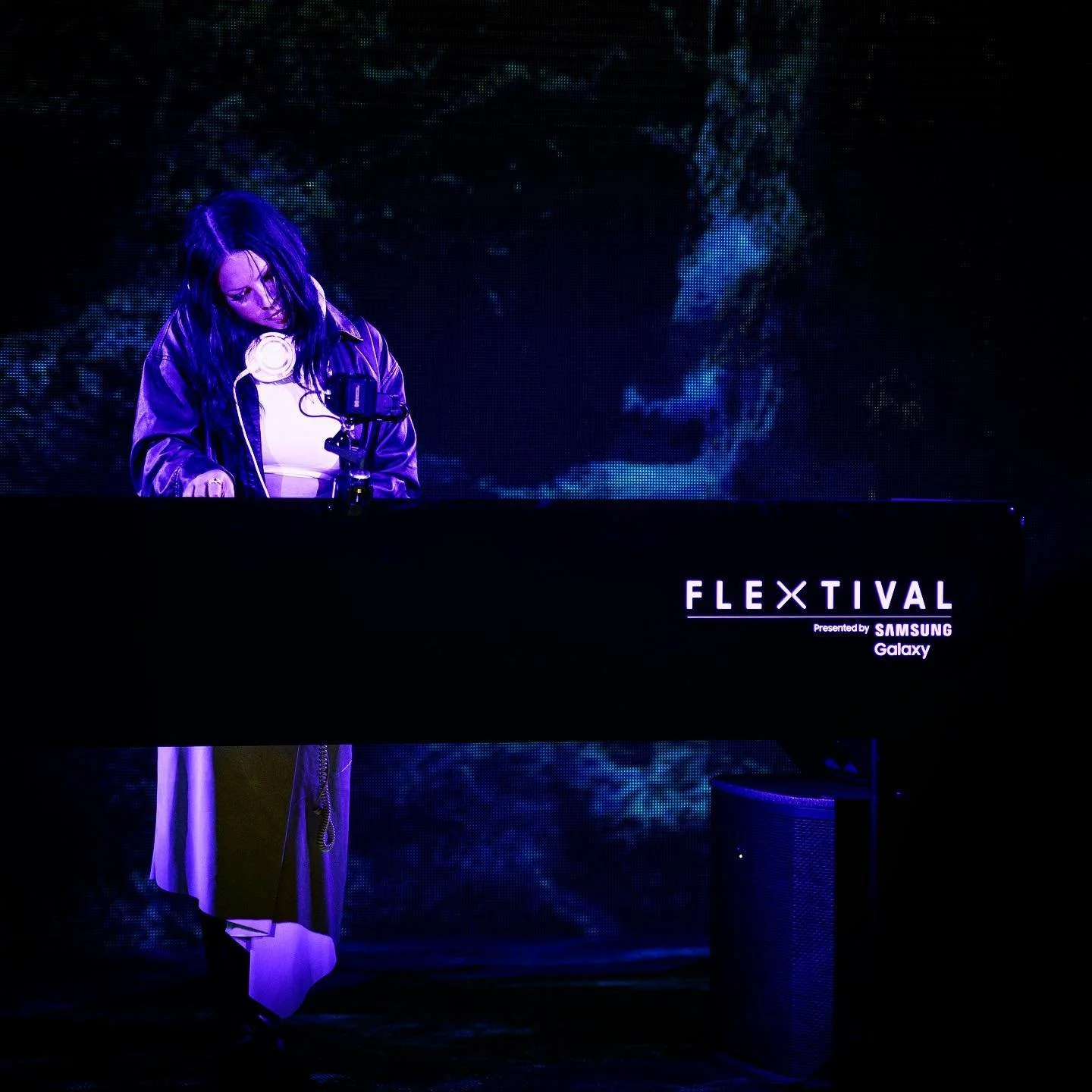 A woman with dark hair, wearing a leather jacket, is DJing at an event called FLEXTIVAL, presented by Samsung Galaxy, with a dark blue and black background.