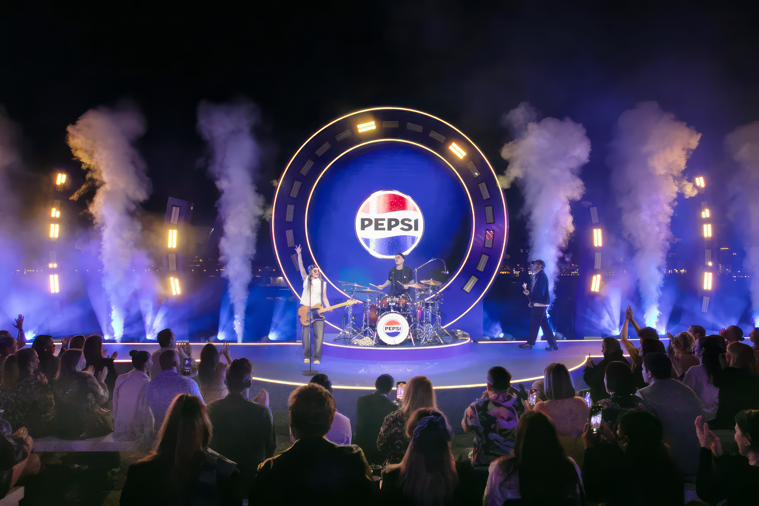 Band performing on stage with Pepsi logo backdrop, surrounded by audience and smoke effects at night.