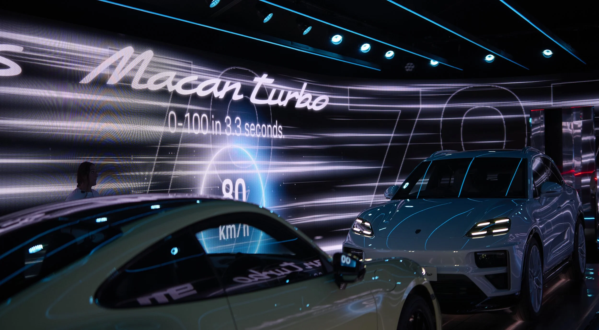 A bright exhibition area featuring a white Macan Turbo car with sleek design and LED headlight details, displayed in front of a large digital screen showcasing car specifications like 0-100 km/h in 3.3 seconds and maximum speed of 280 km/h, with vibr