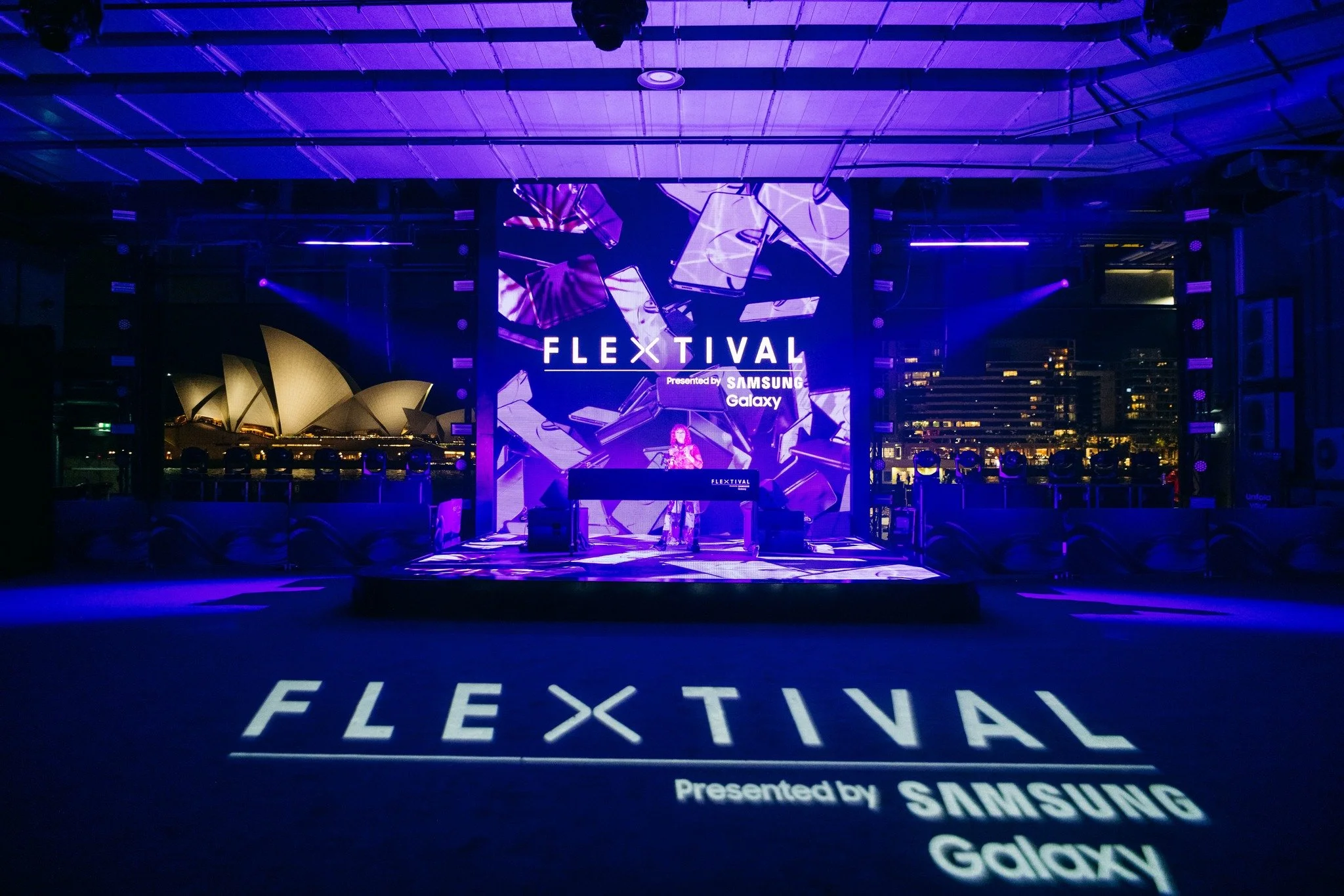 A stage with a large digital screen displaying 'FLEXLIVAL presented by Samsung Galaxy', decorated with purple lighting and graphics of smartphones, with a performer or DJ on stage at night. The floor projects the same 'FLEXLIVAL' branding.