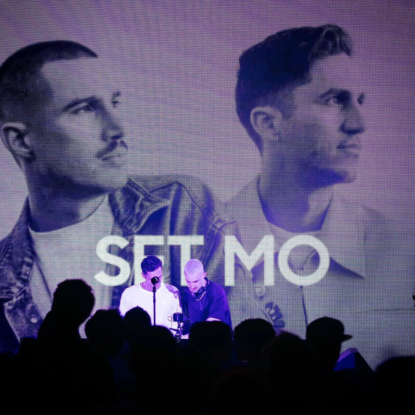 Two men on stage in front of a large black-and-white image of two young men with the word 'SILIMO' displayed below. The stage is dark, with silhouettes of the audience visible.