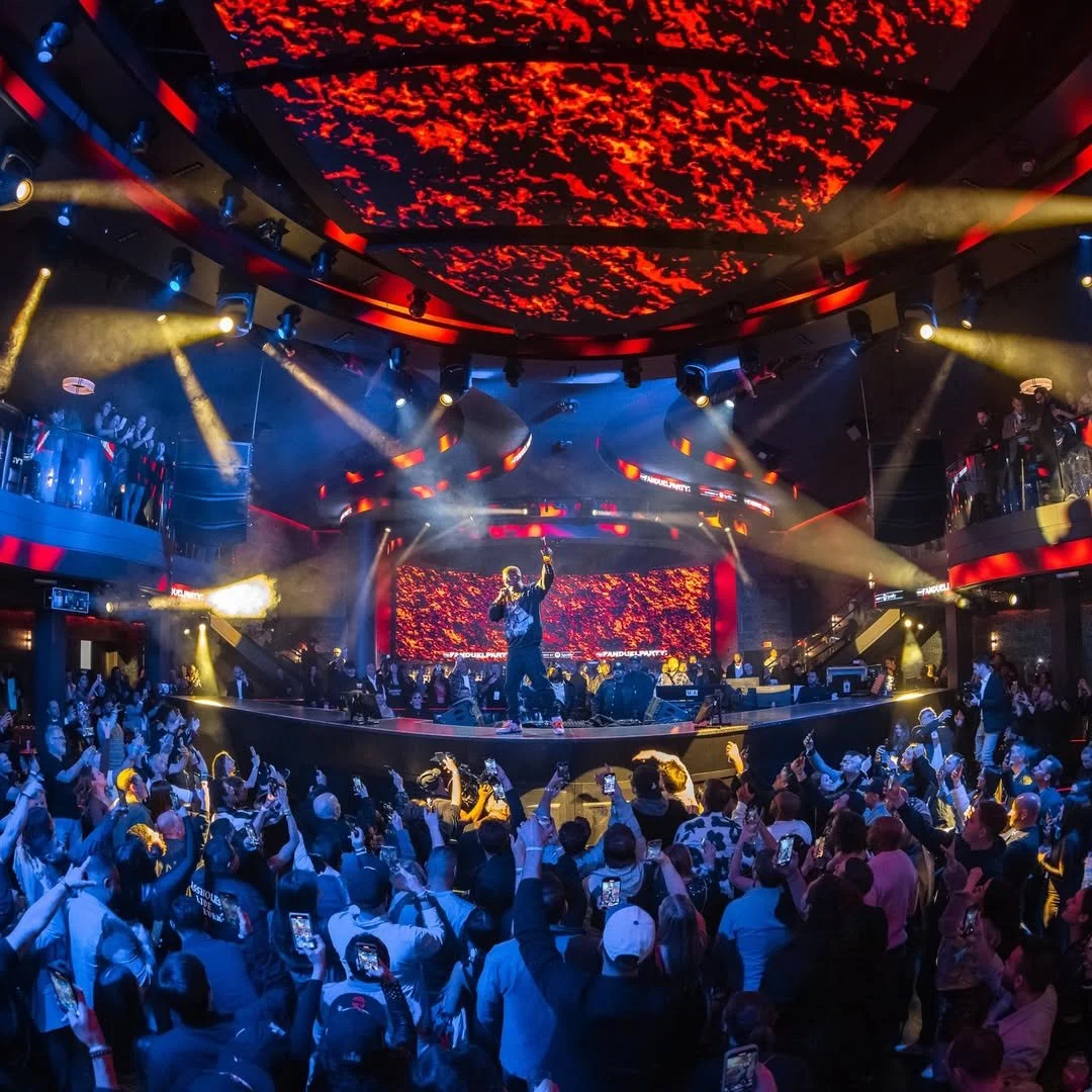 A live concert inside a vibrant nightclub with a crowd facing the stage, performer on stage with a microphone, colorful lighting, and a large LED screen with red and orange visuals overhead.