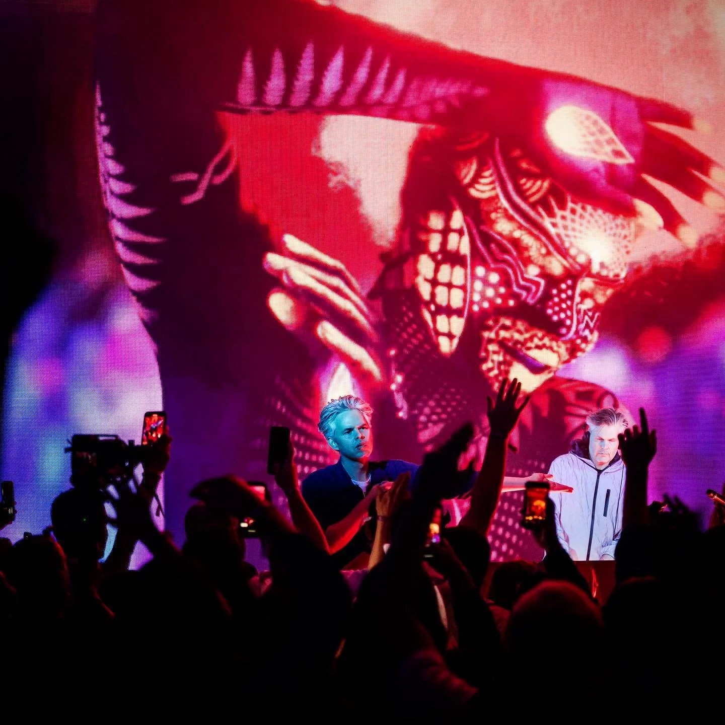 A musical performance with two men on stage in front of a large, colorful digital art screen, with an audience capturing the moment on their phones.