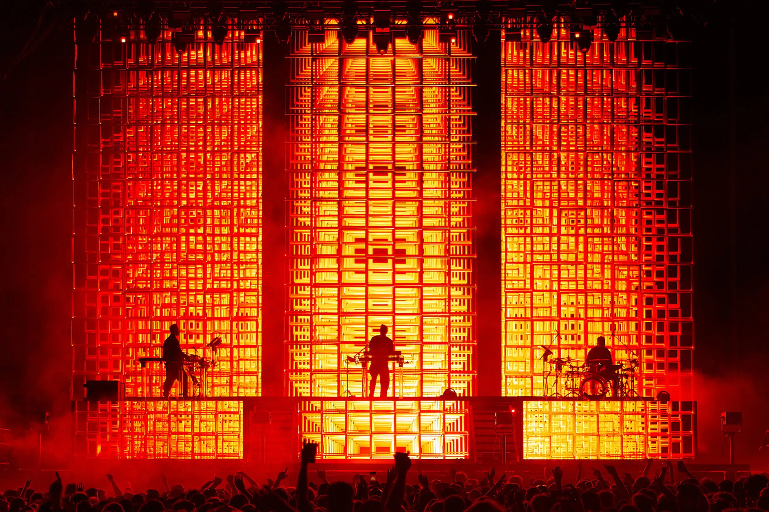 Silhouettes of musicians performing on a stage with a bright orange and red grid backdrop, creating a vibrant and dynamic concert atmosphere.