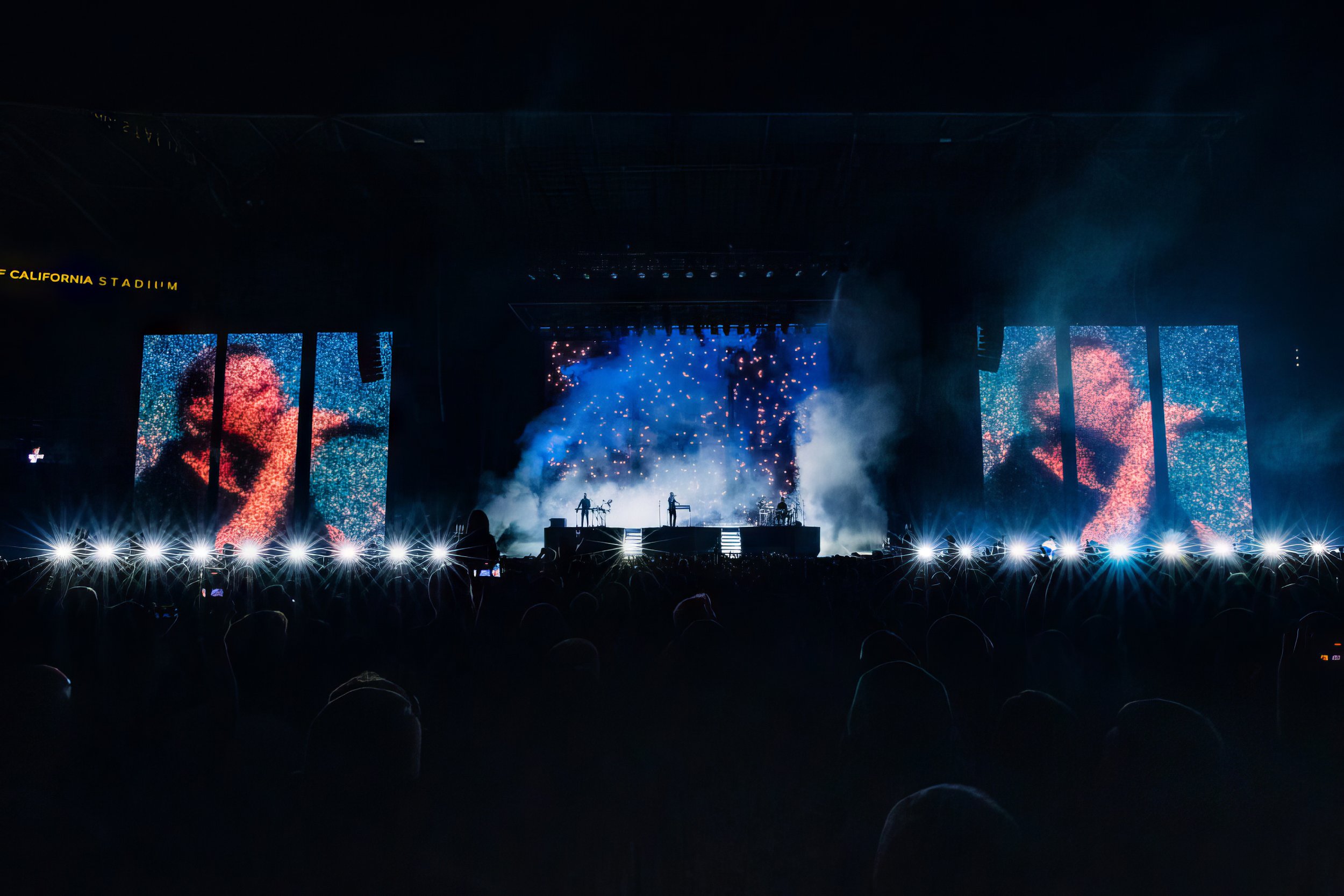 Large concert with bright lights and video screens, featuring a band performing on stage with a background of starry visuals and smoke effects.