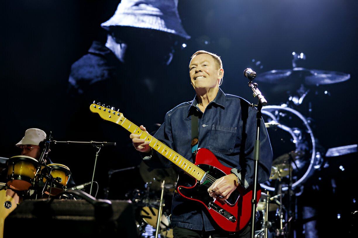Musician playing electric guitar on stage with a band, including a percussionist in the background, and large screen display behind.