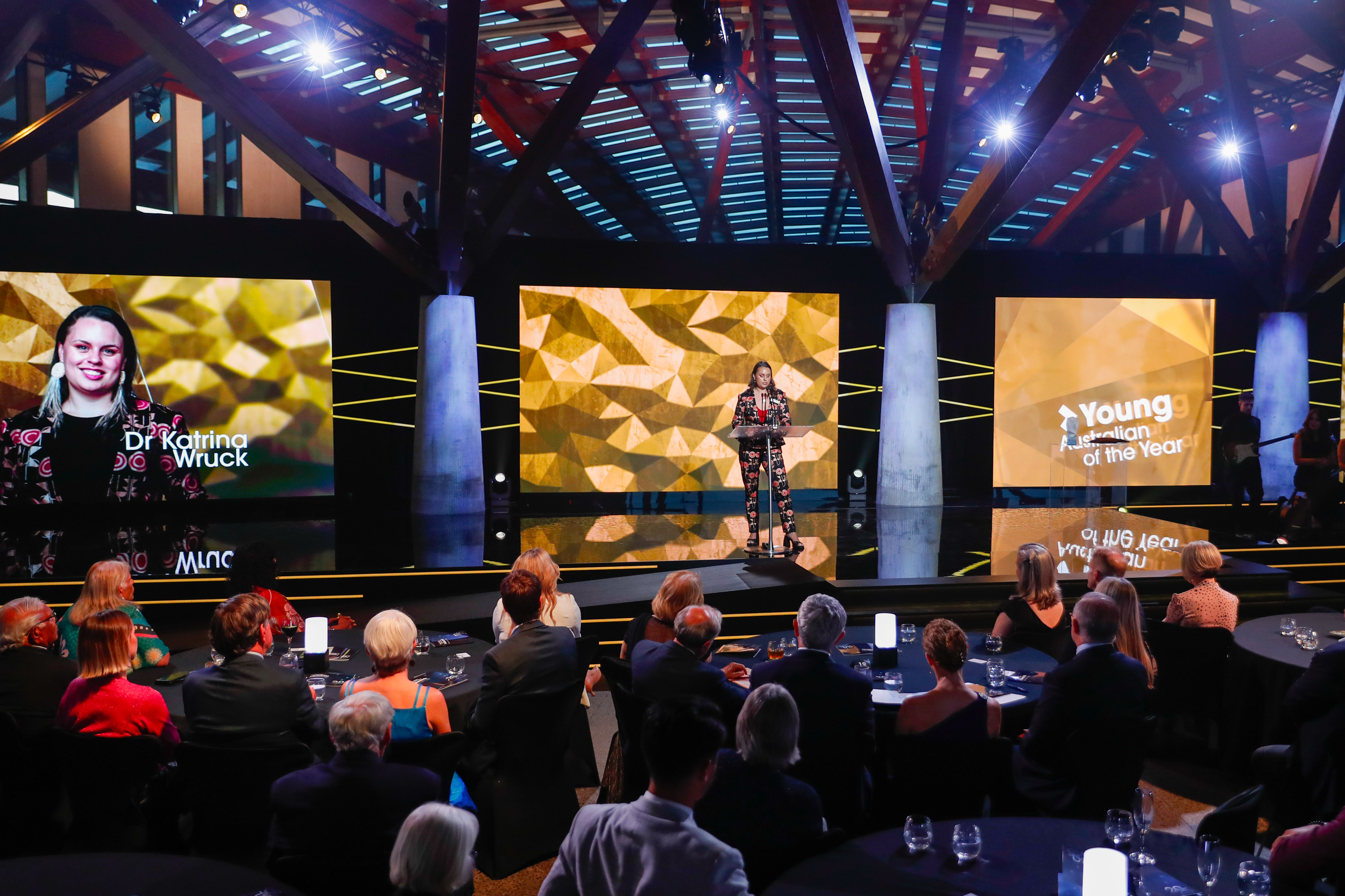 An award ceremony on a stage with a large audience seated at round tables. A woman is speaking at a podium, with three large screens behind her displaying gold geometric backgrounds. The screen on the left shows a woman's portrait with the text "Dr K