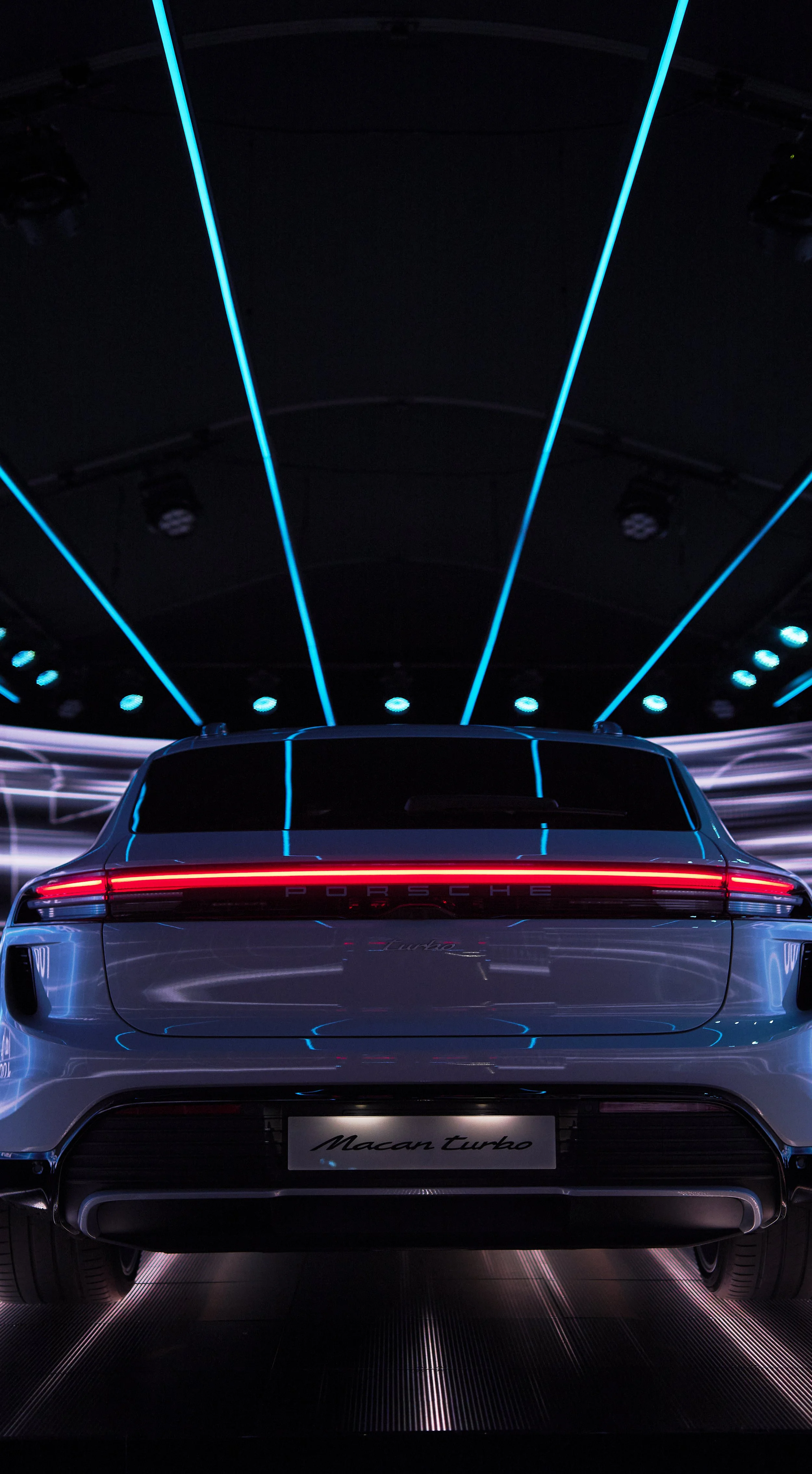 Rear view of a silver Porsche Macan Turbo with distinctive red light bar across the back, illuminated blue and white ceiling lights, and neon accents, in a dark, modern showroom.