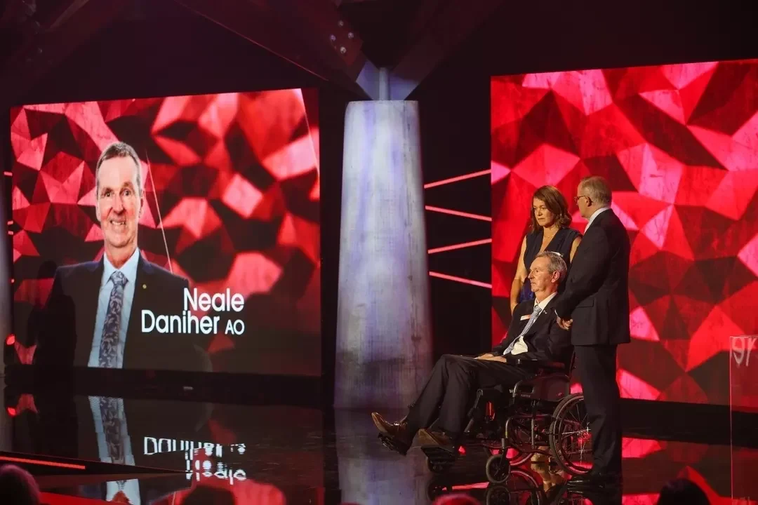Neale Daniher, an AFL football legend, seated in a wheelchair on stage during an awards event, with two people standing beside him and a large screen behind displaying his name and image against a red geometric background.