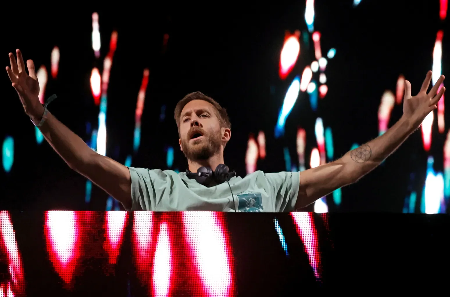 A male DJ with arms outstretched, wearing headphones around his neck, standing behind a DJ booth with colorful lights and abstract light patterns in the background.