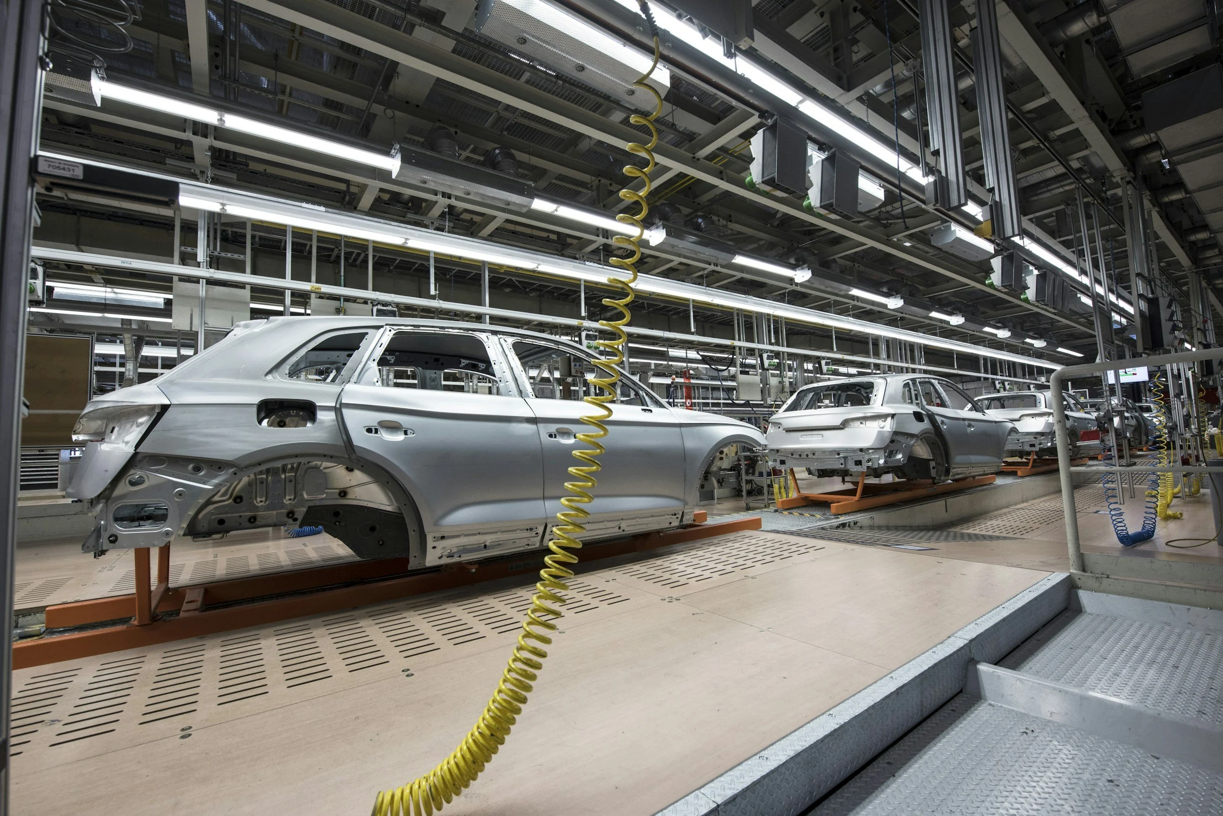 Automobile assembly line with several silver car bodies in various stages of production inside a factory.
