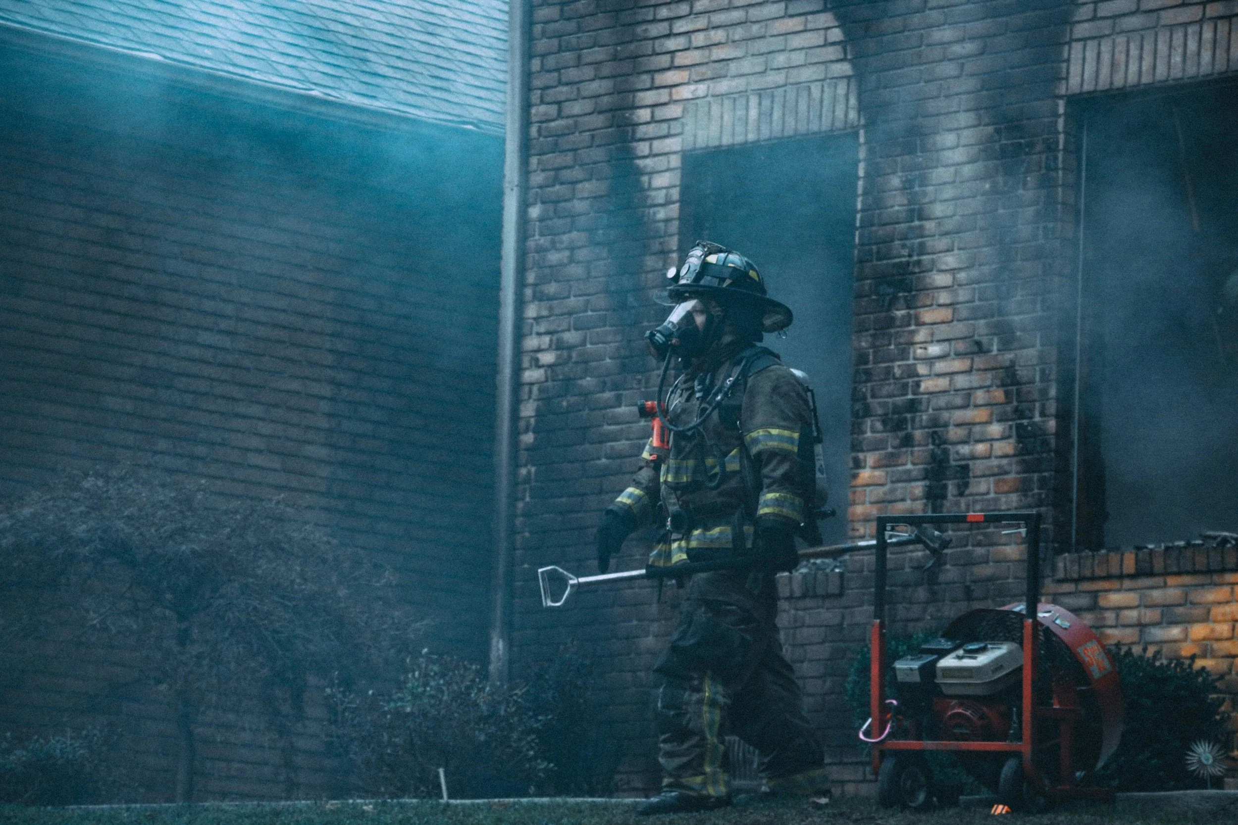 Firefighter in full gear holding a tool near a fire-damaged brick building with smoke and soot on the wall.