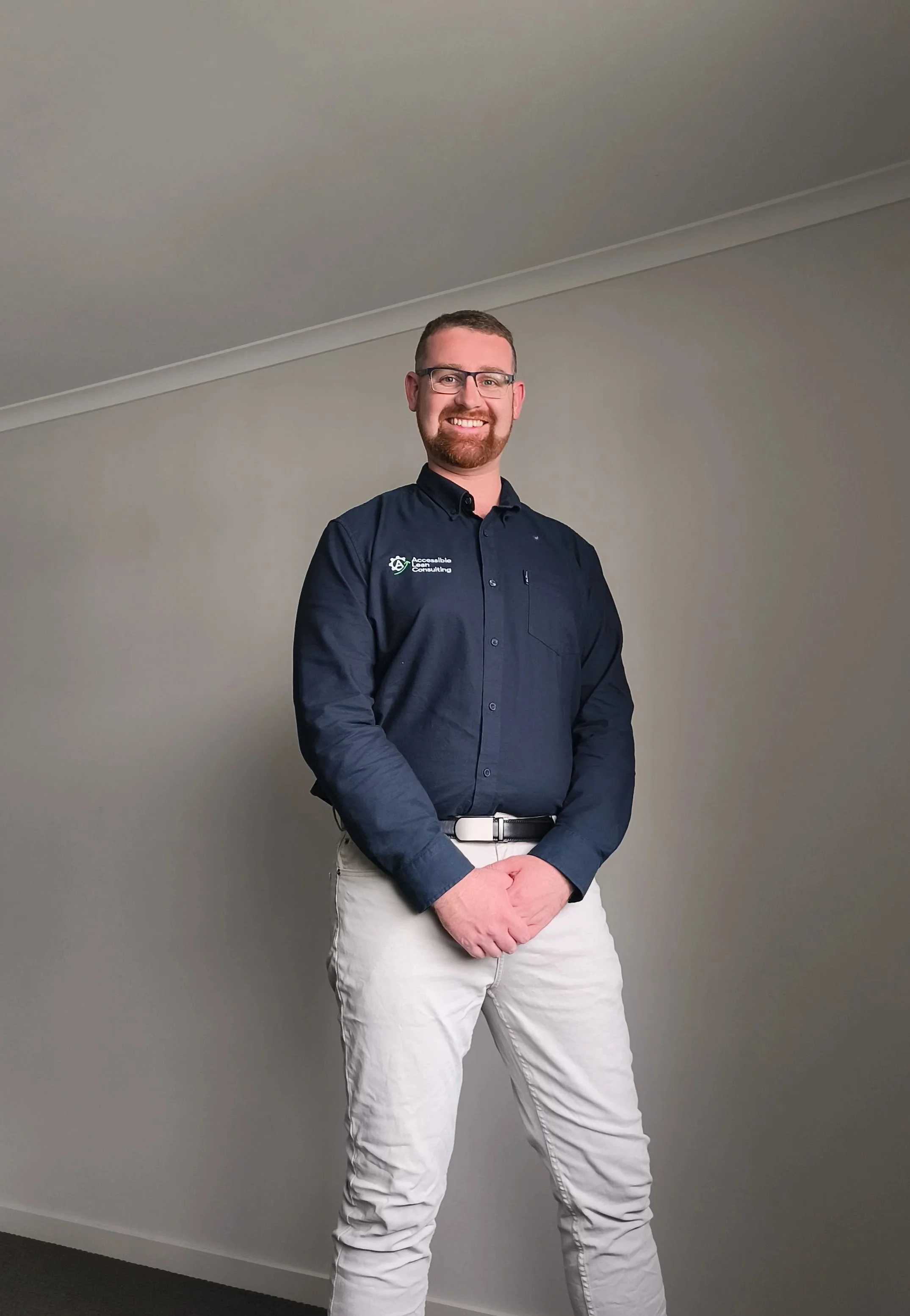 A smiling man with glasses, beard, and short hair wearing a navy blue shirt with a logo that reads 'Accessible Consulting' and beige pants, standing against a plain gray wall.