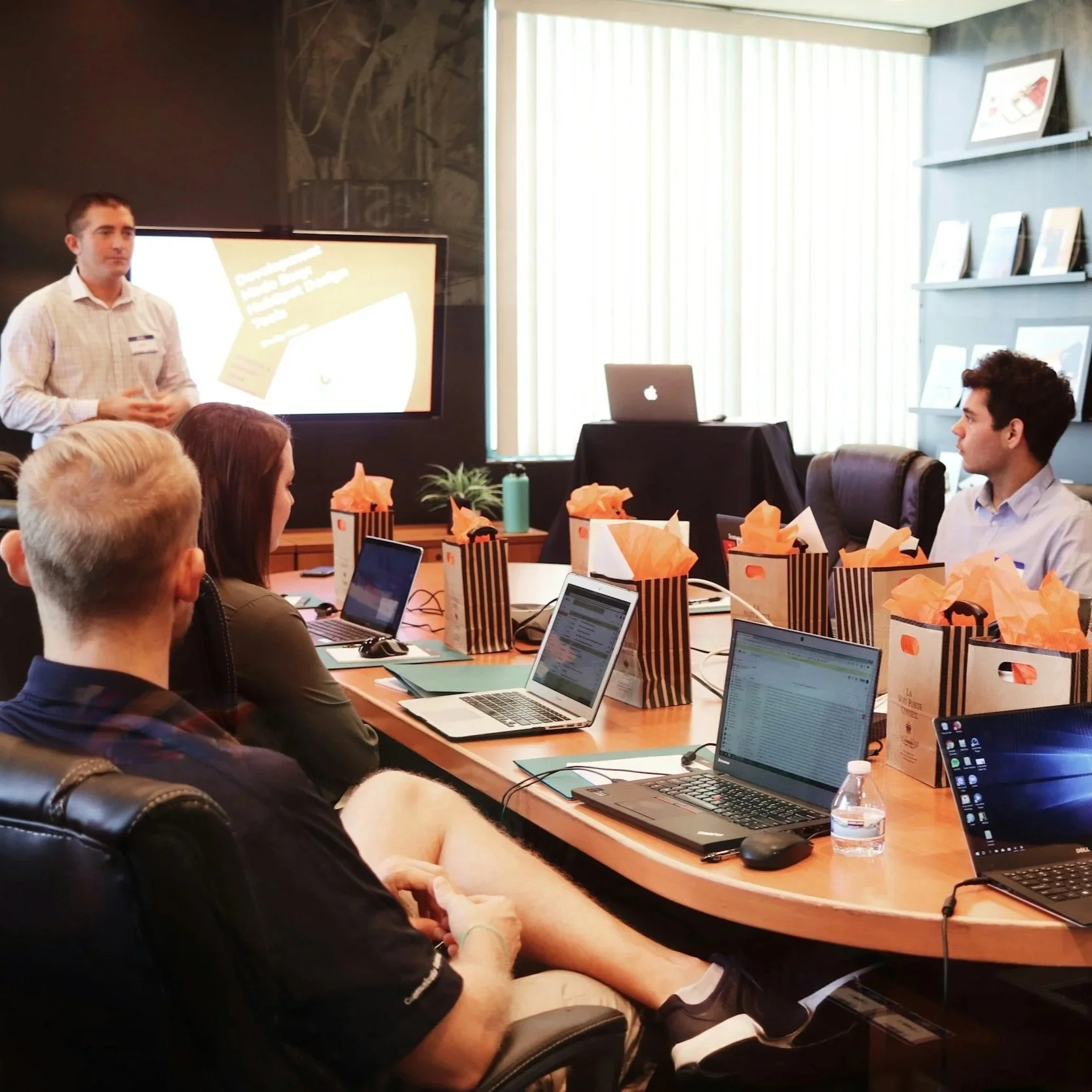 A man is standing at the front of a conference room giving a presentation to seated attendees. The room has laptops and gift bags on the table, and a large screen displaying a slide.