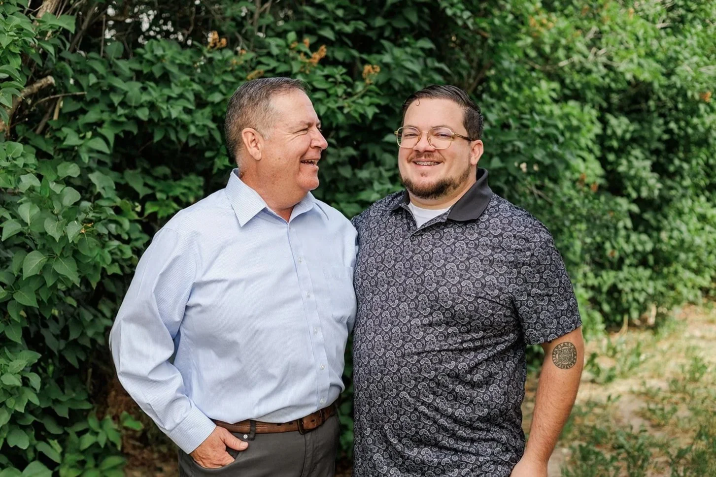 Two men standing outdoors, smiling and looking at each other, with green foliage in the background.