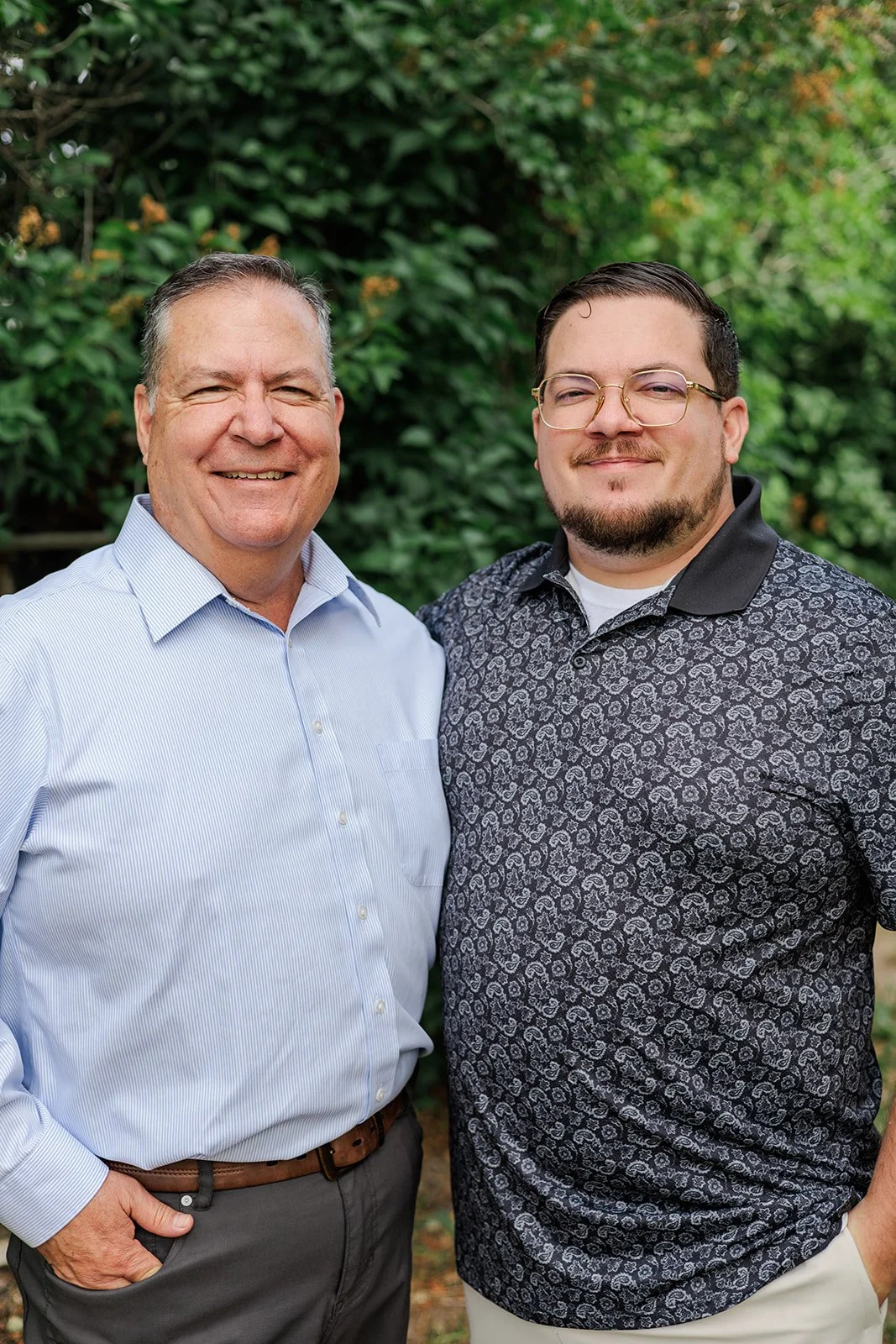 Two men standing close together outdoors in front of greenery, smiling at the camera. The man on the left is older with short gray hair, wearing a light blue button-up shirt and dark pants. The man on the right is younger with glasses, a beard, and short dark hair, wearing a black patterned polo shirt and light-colored pants.