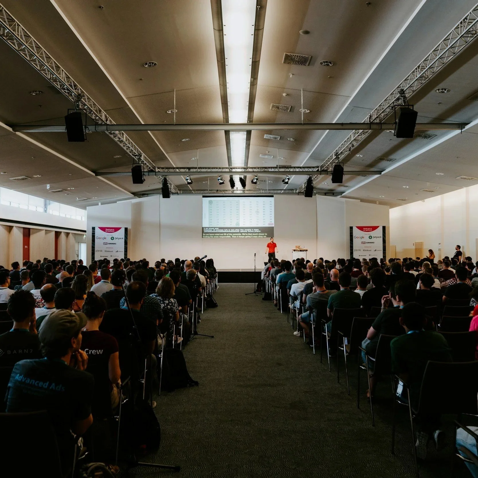 A large conference room filled with a seated audience listening to a presenter on stage. The stage has a large screen displaying a chart or data, and there are banners and lighting equipment overhead.
