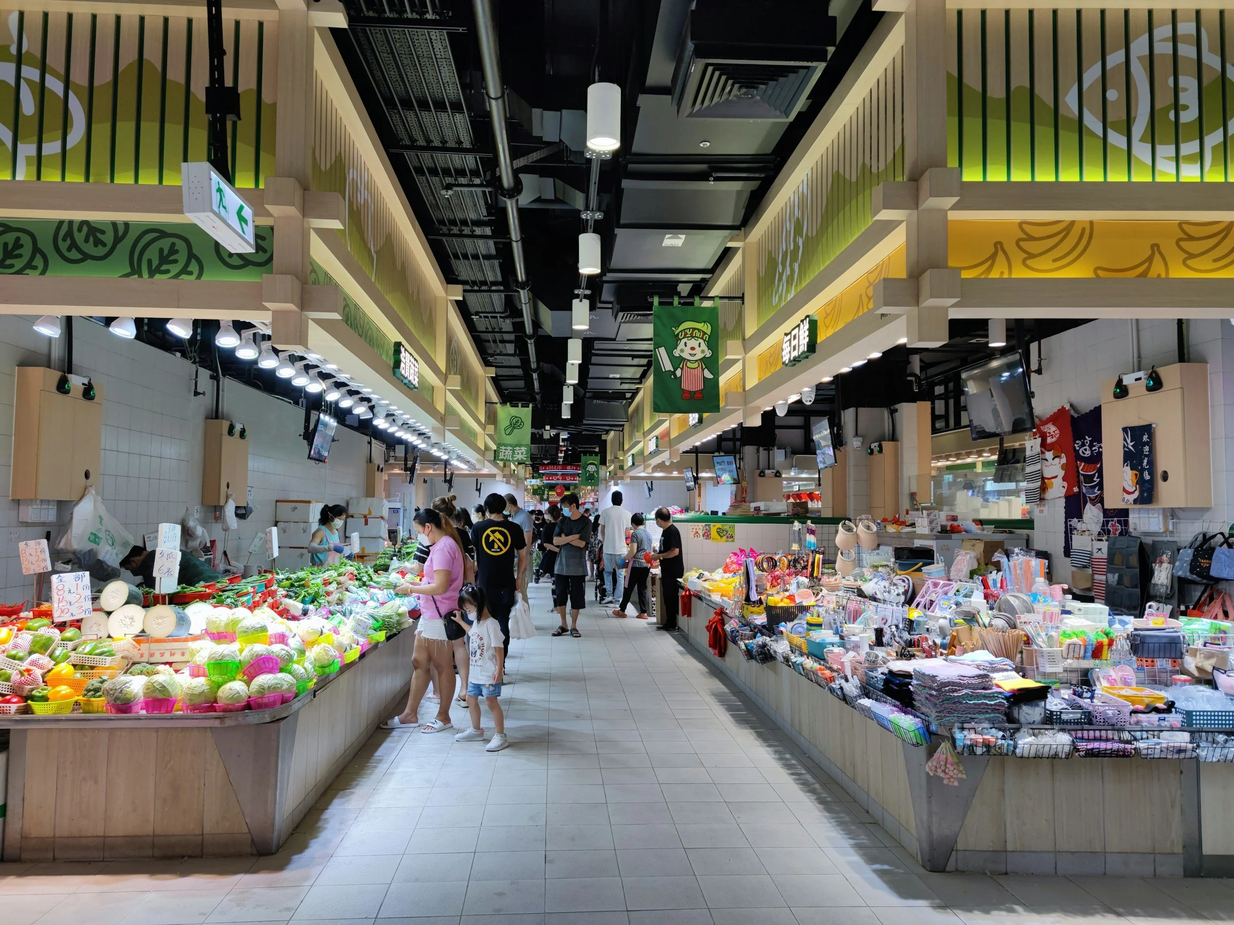 Vista interior de un mercado con puestos de frutas, verduras y artículos diversos. Hay varias personas comprando y caminando al fondo.